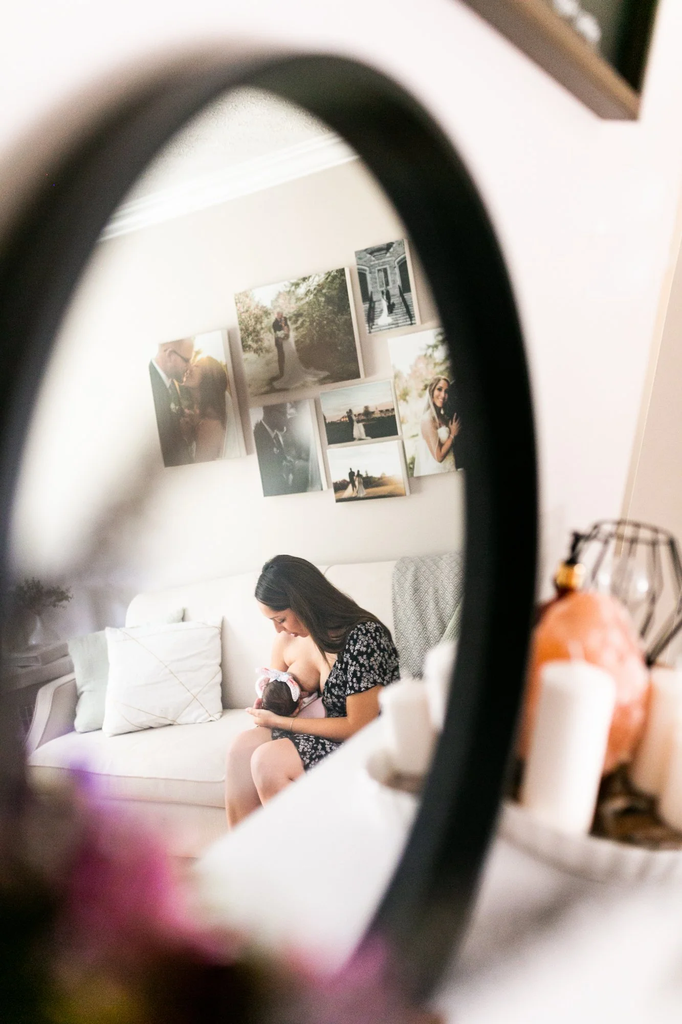 Woman breastfeeding infant on couch, reflected in a mirror, with photo collage on wall.