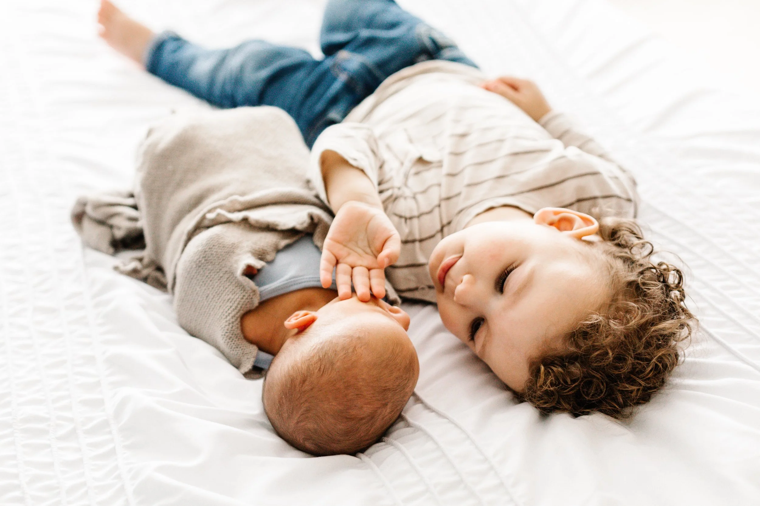 Young child laying beside a newborn baby on a white bed, sibling bonding moment.