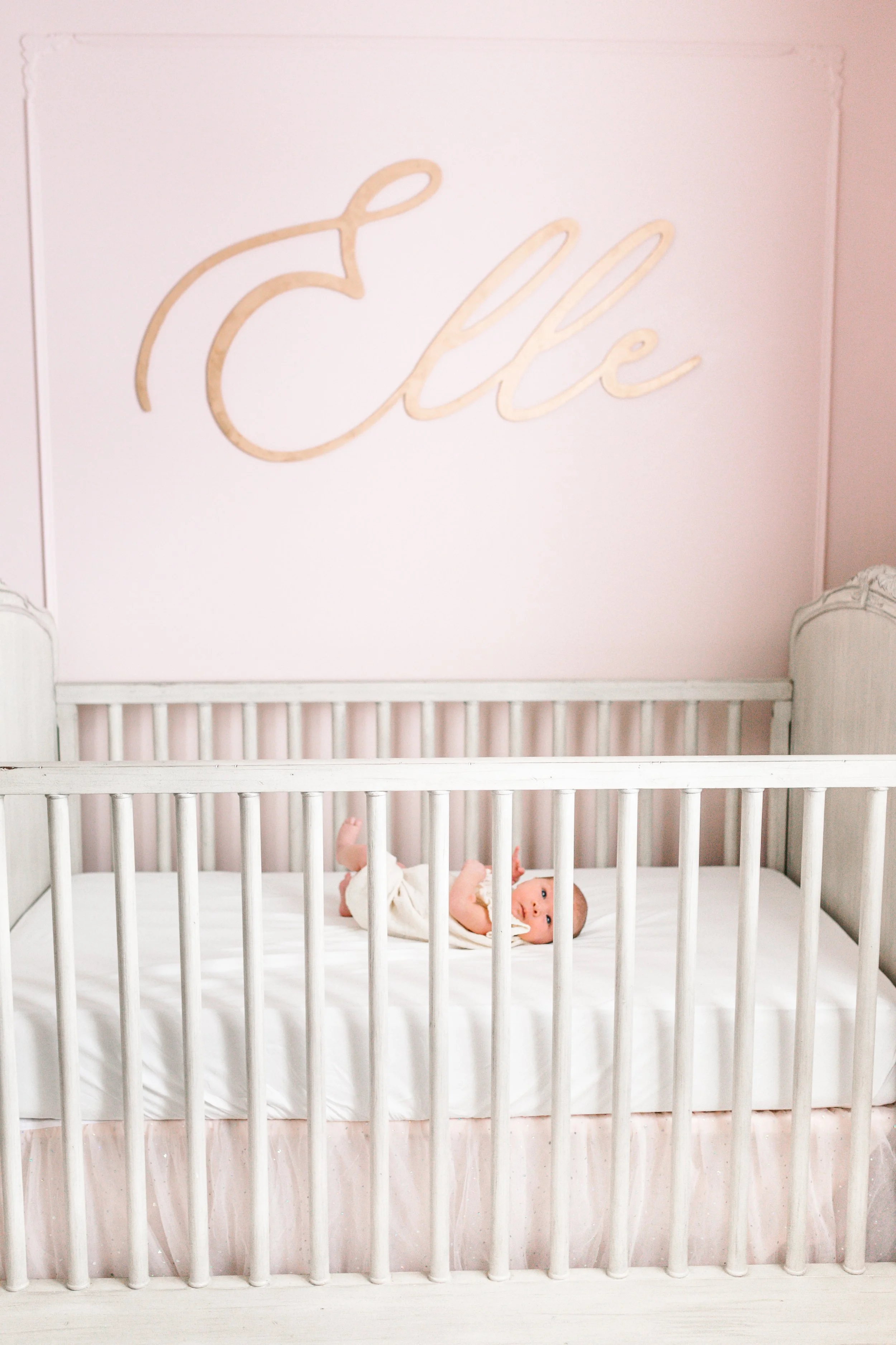 Baby lying in a white crib with a light pink wall and "Elle" written in cursive above.
