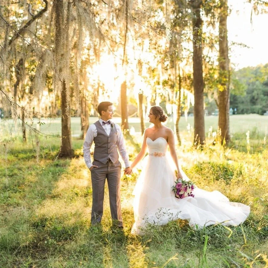A couple standing in a sunlit forest wearing wedding attire, holding hands and smiling at each other, with the bride holding a bouquet.