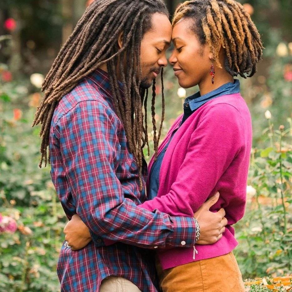 A couple embracing gently in a garden, with their foreheads touching and eyes closed. The man is wearing a plaid shirt, and the woman is wearing a magenta sweater.