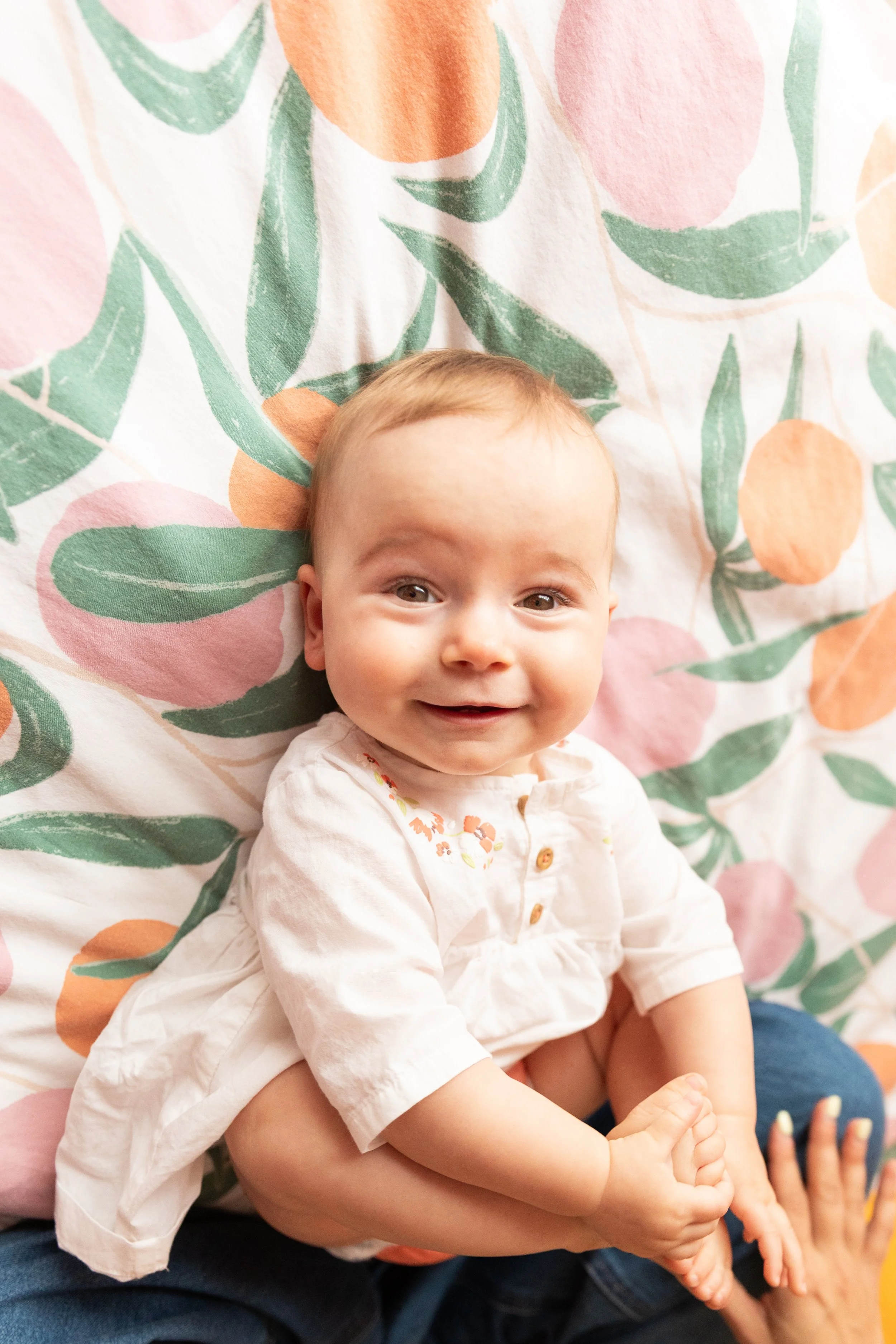 A smiling baby with light skin and red hair, wearing a white shirt, sitting on a person's lap, with a colorful floral background.