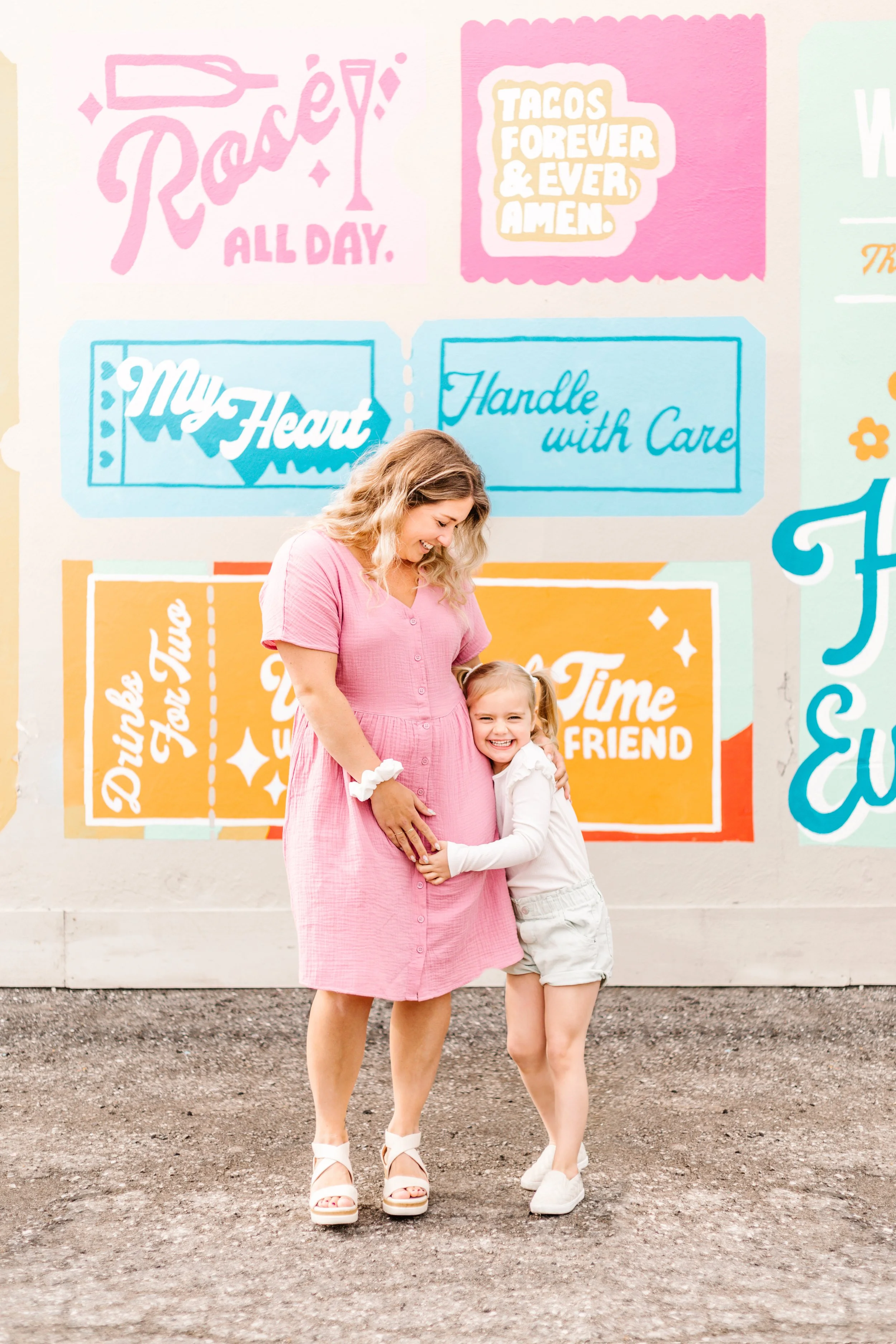 A woman in a pink dress and a child hugging her, standing in front of a colorful wall with fun text like "Rosé All Day" and "Tacos Forever & Ever, Amen."