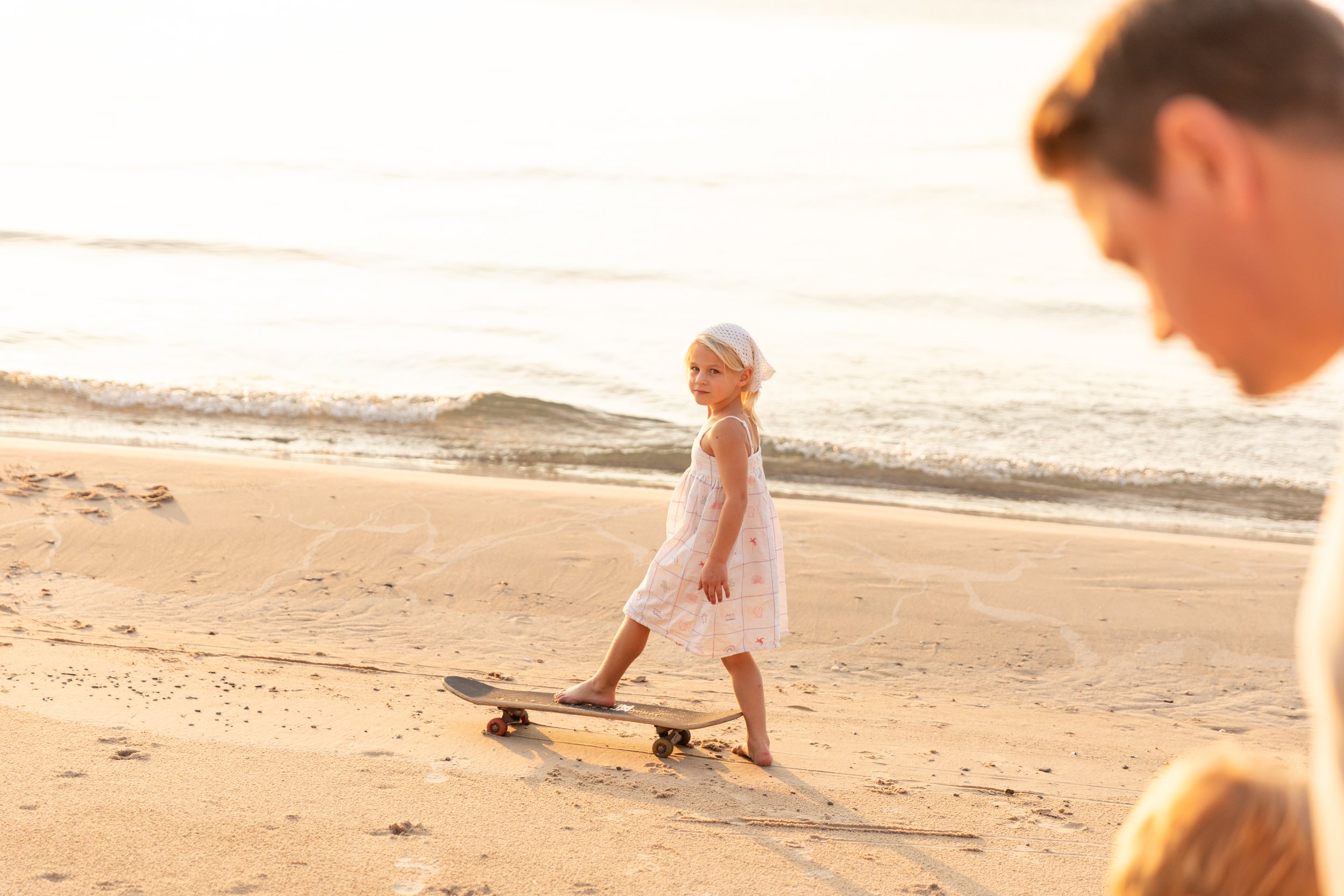 A young girl in a light-colored dress and headscarf standing on a skateboard on a sandy beach near the water, with a man and possibly another child partially visible in the foreground.