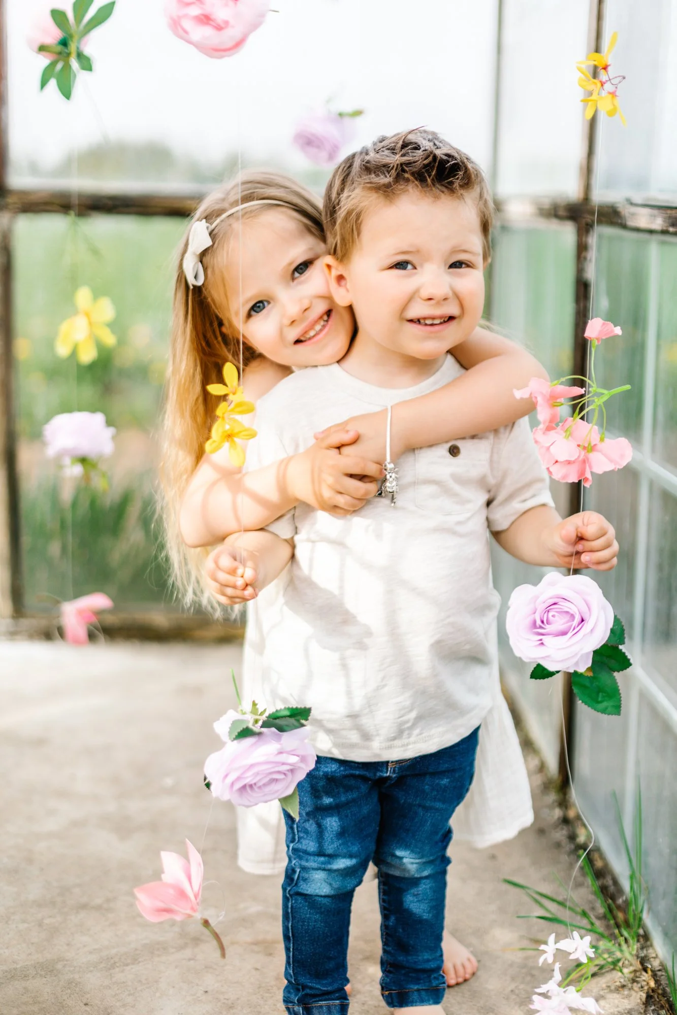 Two young children, a girl and a boy, standing together indoors surrounded by colorful hanging flowers, smiling and hugging each other.