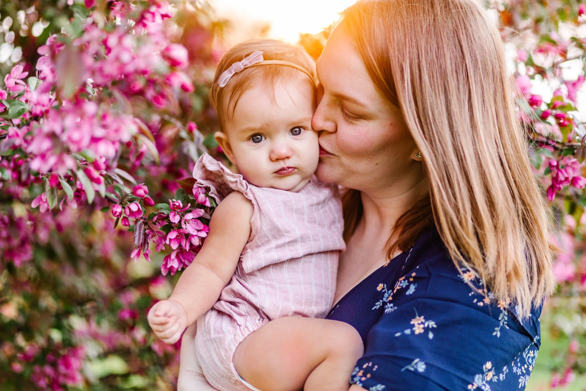 Woman kissing baby near pink flowering tree.