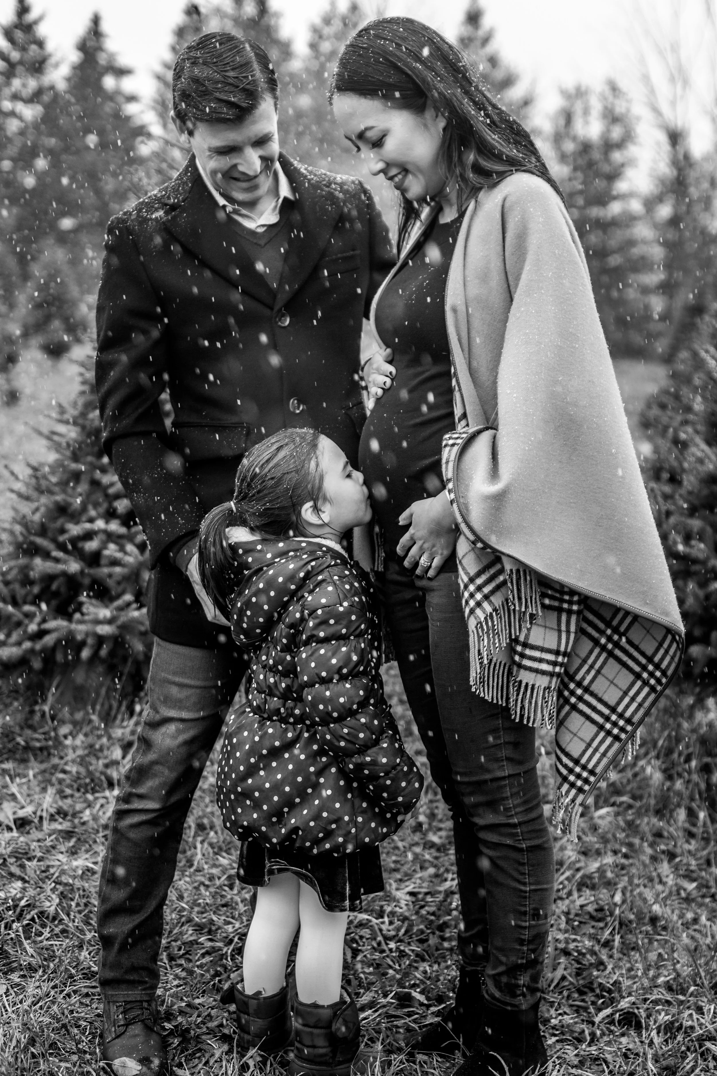 A black and white photo of a family outdoors in winter. A woman, visibly pregnant, is standing with a man and a child. The child is kissing the pregnant belly while snow falls around them.