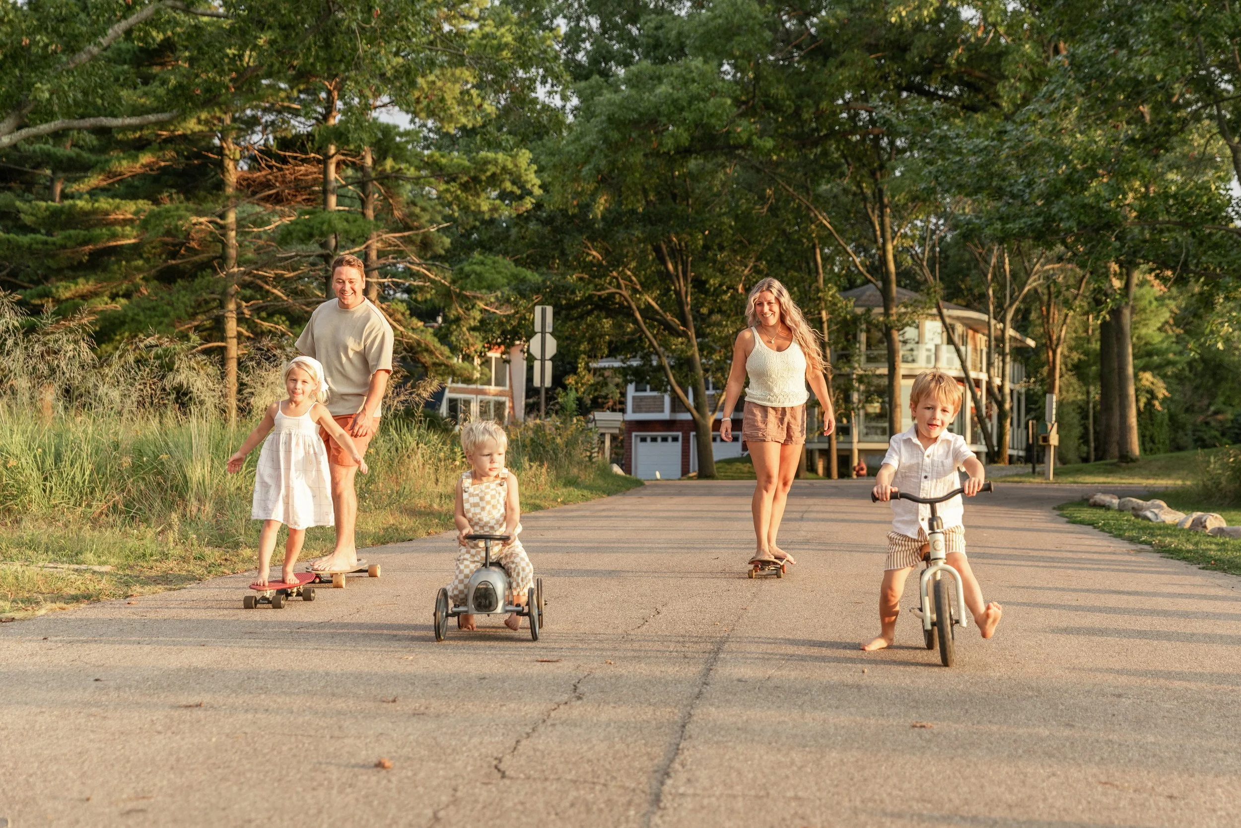 A family of five enjoying outdoor activities on a sunny evening, with children riding bikes and toddlers on tricycles, on a neighborhood street lined with trees and houses.