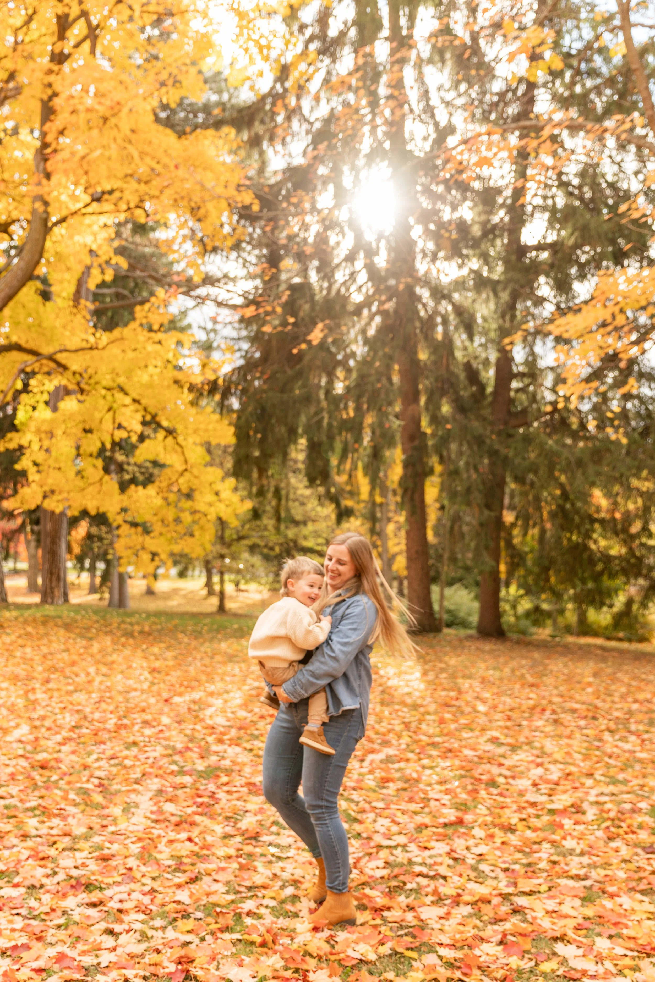 A woman and a young boy are playing together in a park during autumn, surrounded by colorful fall leaves and tall trees, with sunlight shining through the branches.