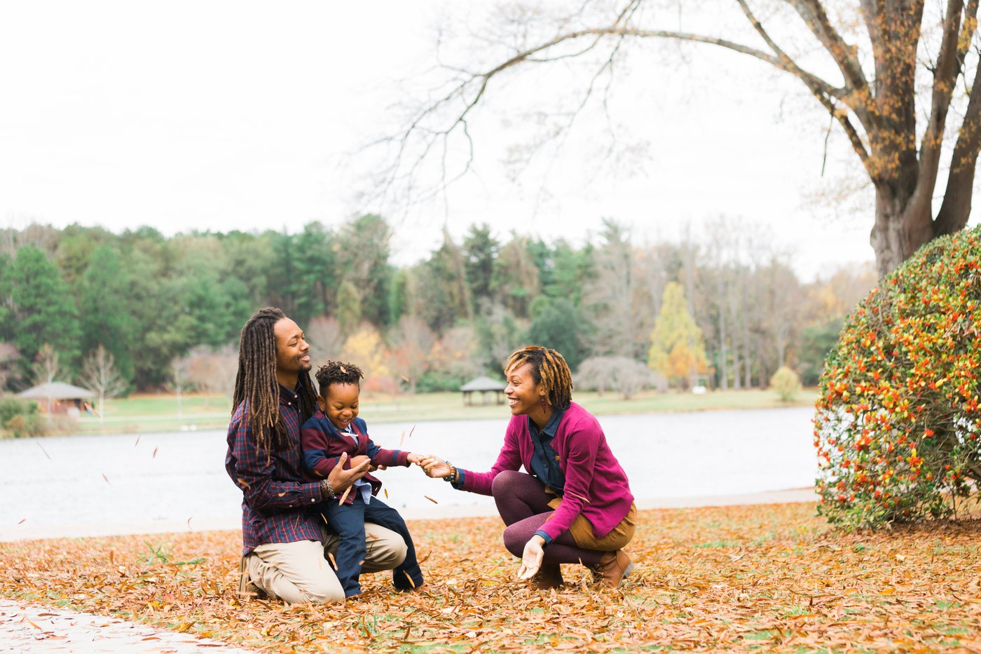 A family of three playing with fallen leaves by a lake in a park during autumn. The parents are smiling and kneeling, while the child is in the father's lap. Trees and shrubs with autumn foliage are in the background.