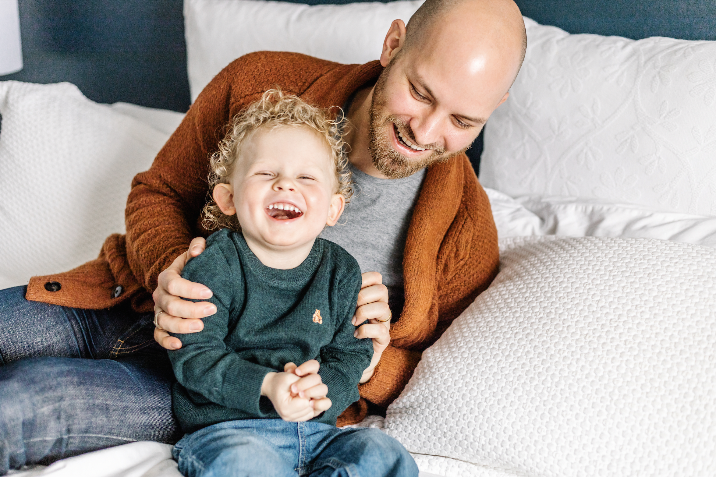 A man and a young boy sitting on a bed, both smiling and laughing. The man is wearing a brown cardigan and the boy is wearing a green sweater. They are surrounded by white pillows and bedding.