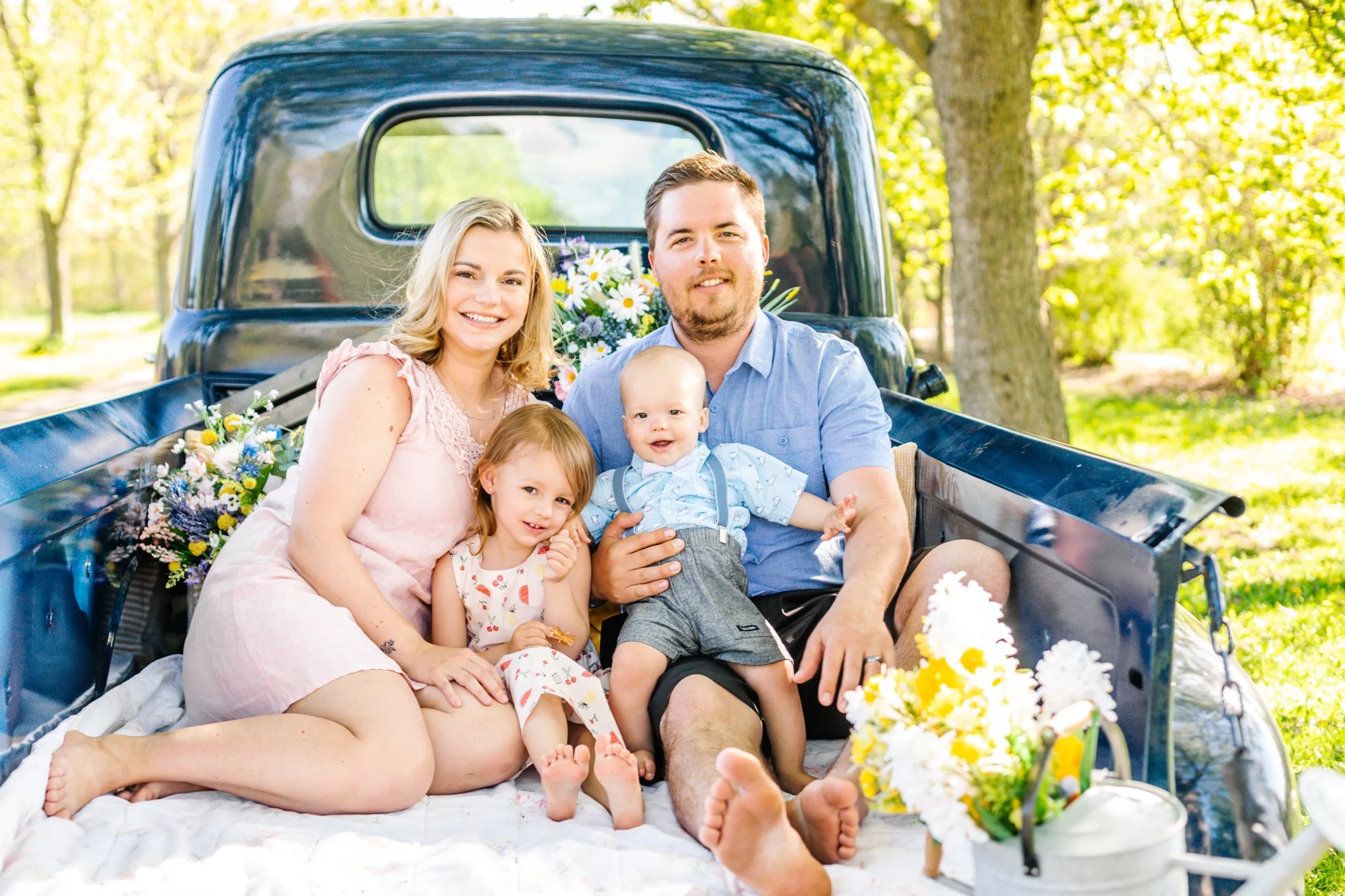Family sitting in the back of a vintage truck, surrounded by flowers, in a park.