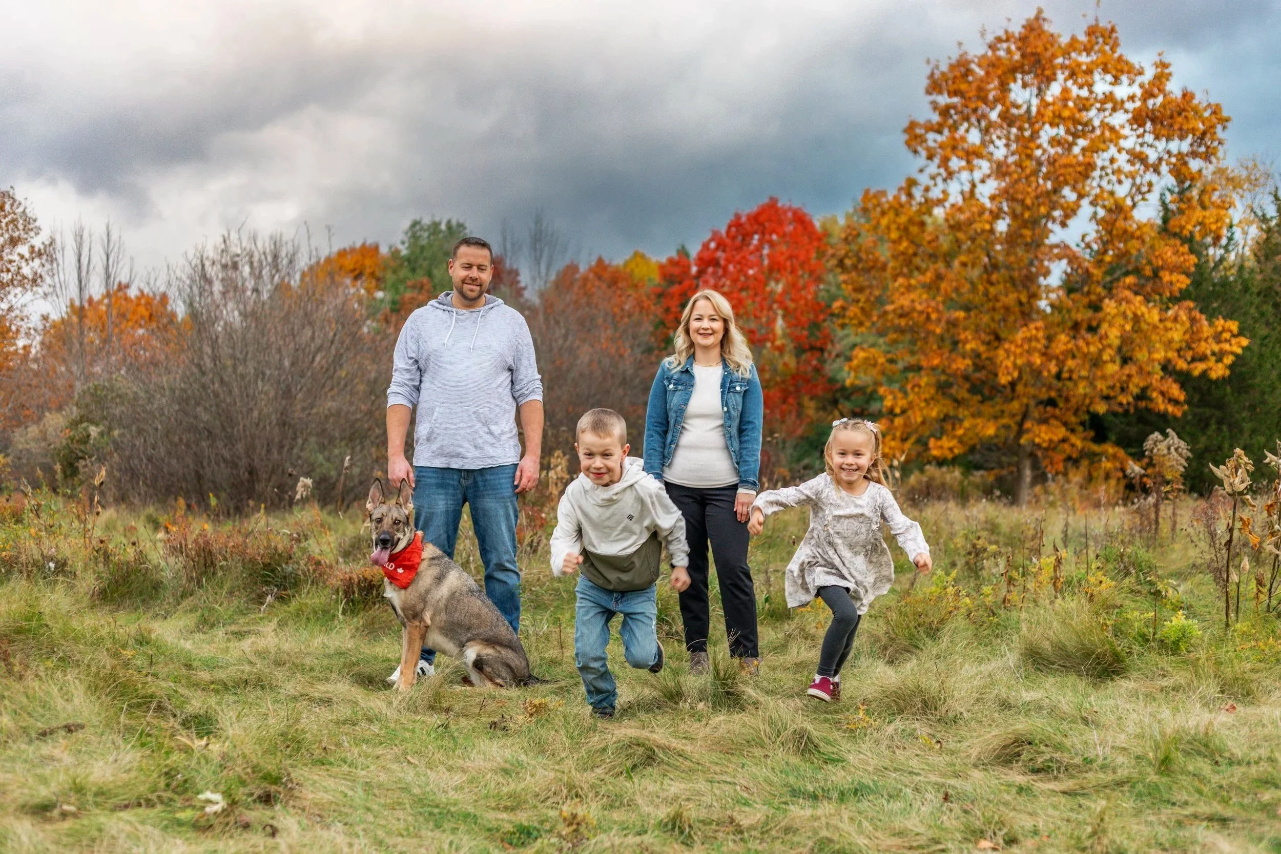 A family of four with a dog poses outdoors in an autumn setting. The parents stand in the background while two children play in the foreground, surrounded by colorful fall foliage.