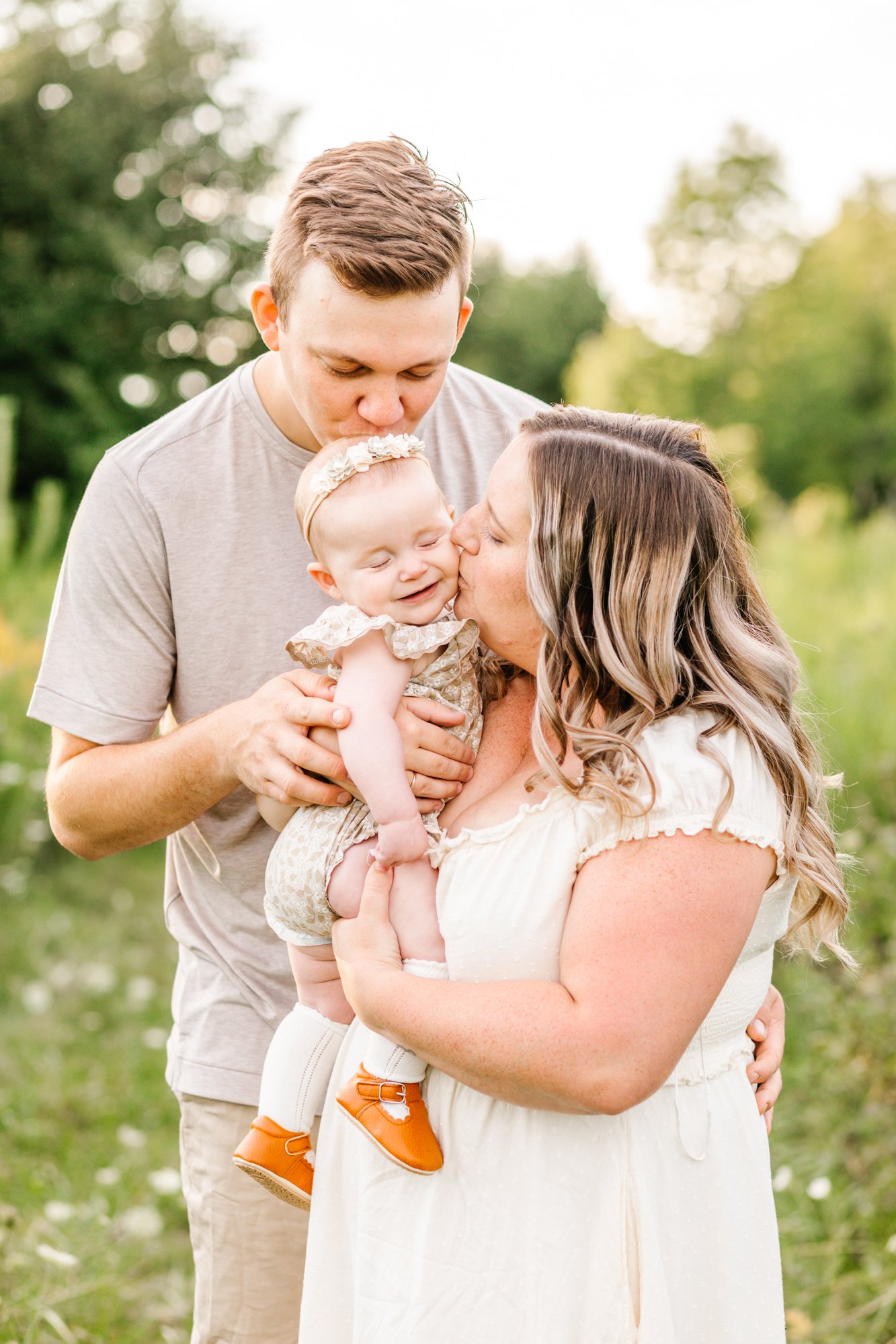 A couple kisses their smiling baby while standing outdoors in a grassy area.