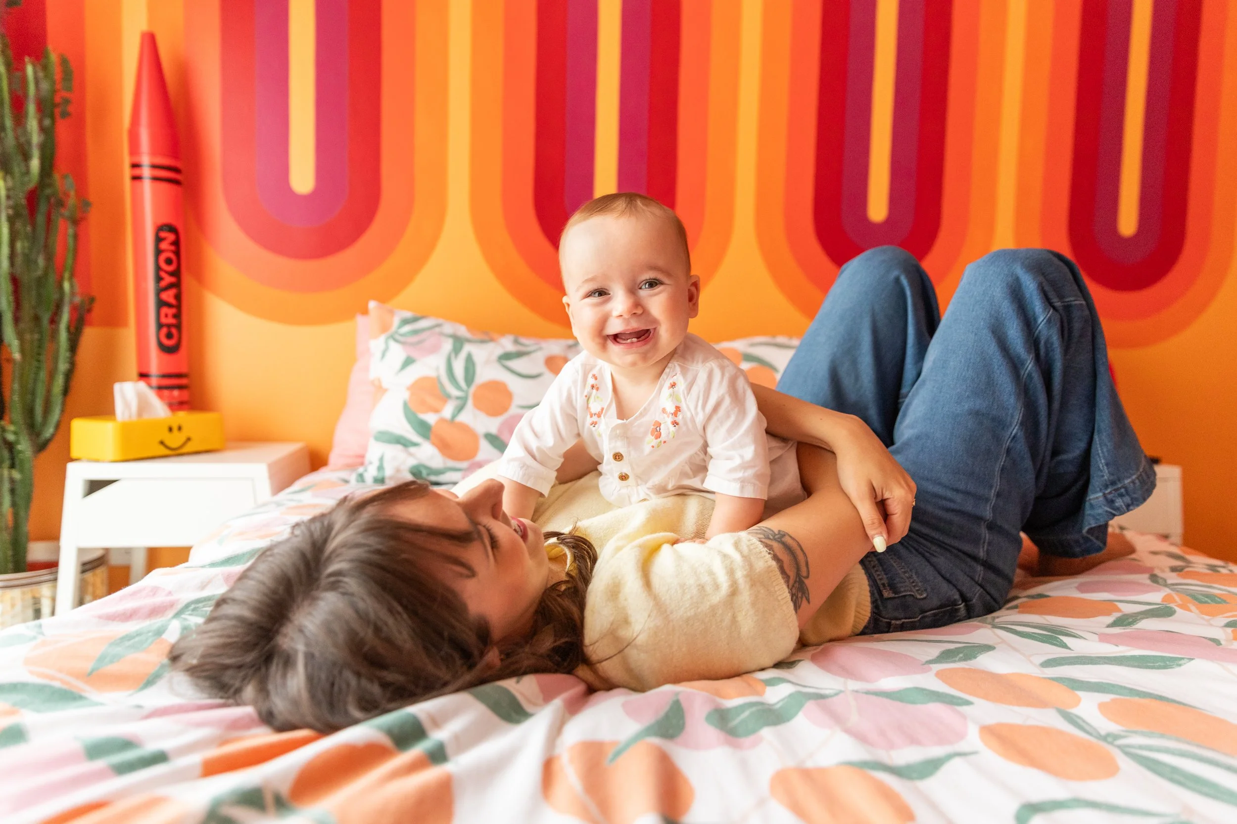 A woman and a small child playing on a bed with a colorful orange and pink patterned wall in the background. The woman is lying on her back, smiling, while the child, smiling , is sitting on her stomach.
