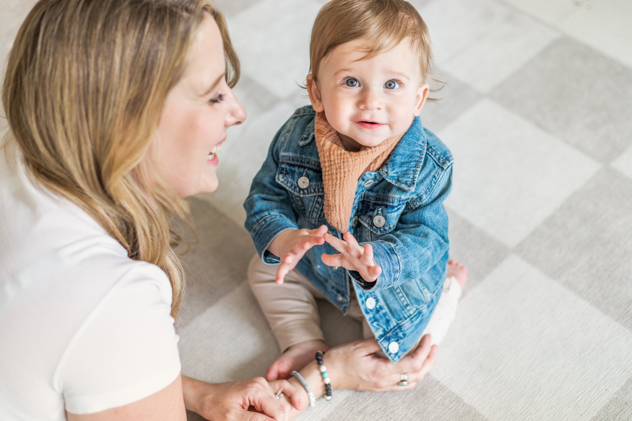 A woman and a young child sitting on a tiled floor, smiling and looking at each other. The child is wearing a denim jacket, beige pants, and a brown scarf.