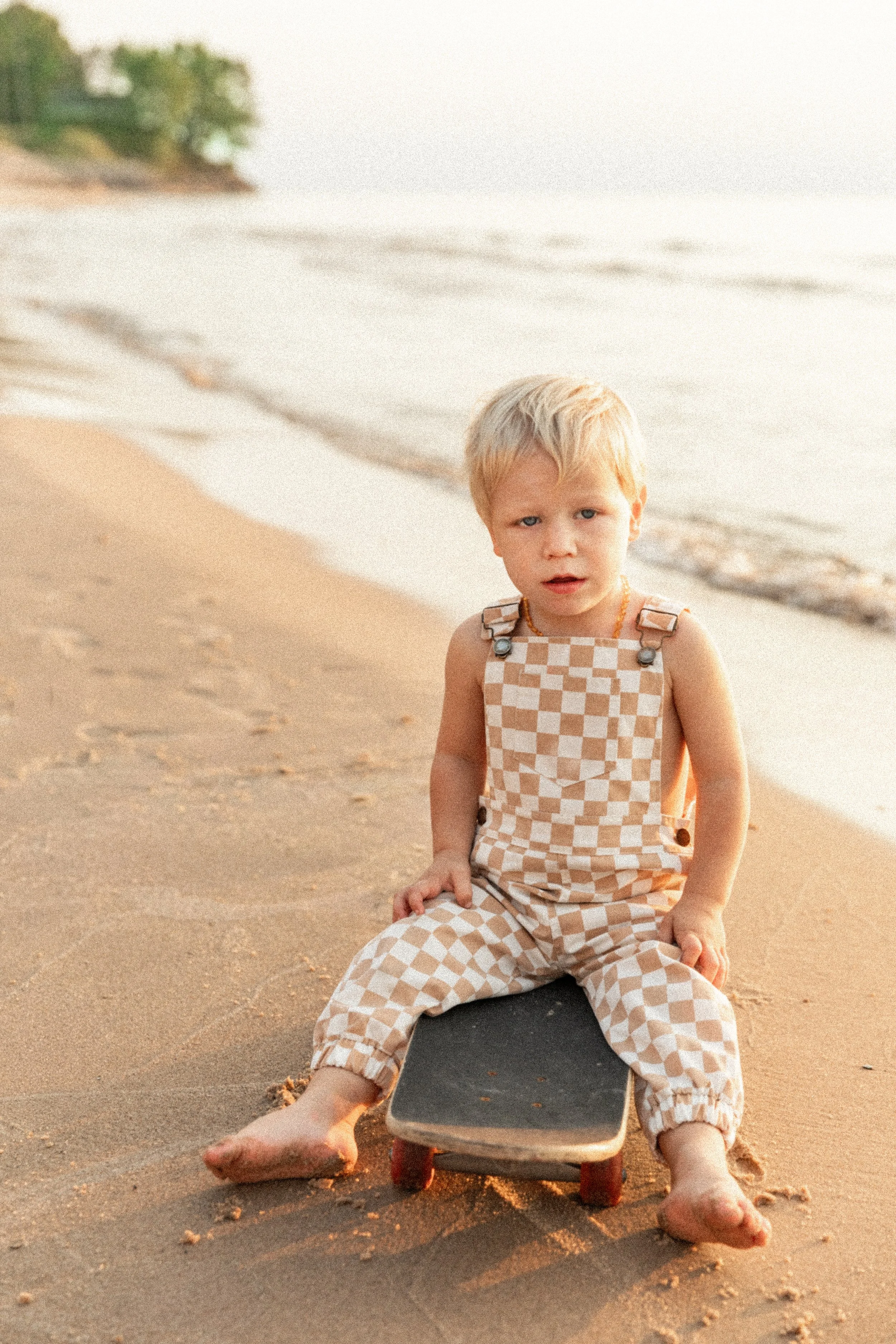 A young blonde boy wearing checkered overalls sitting on a skateboard on a sandy beach with the ocean in the background.