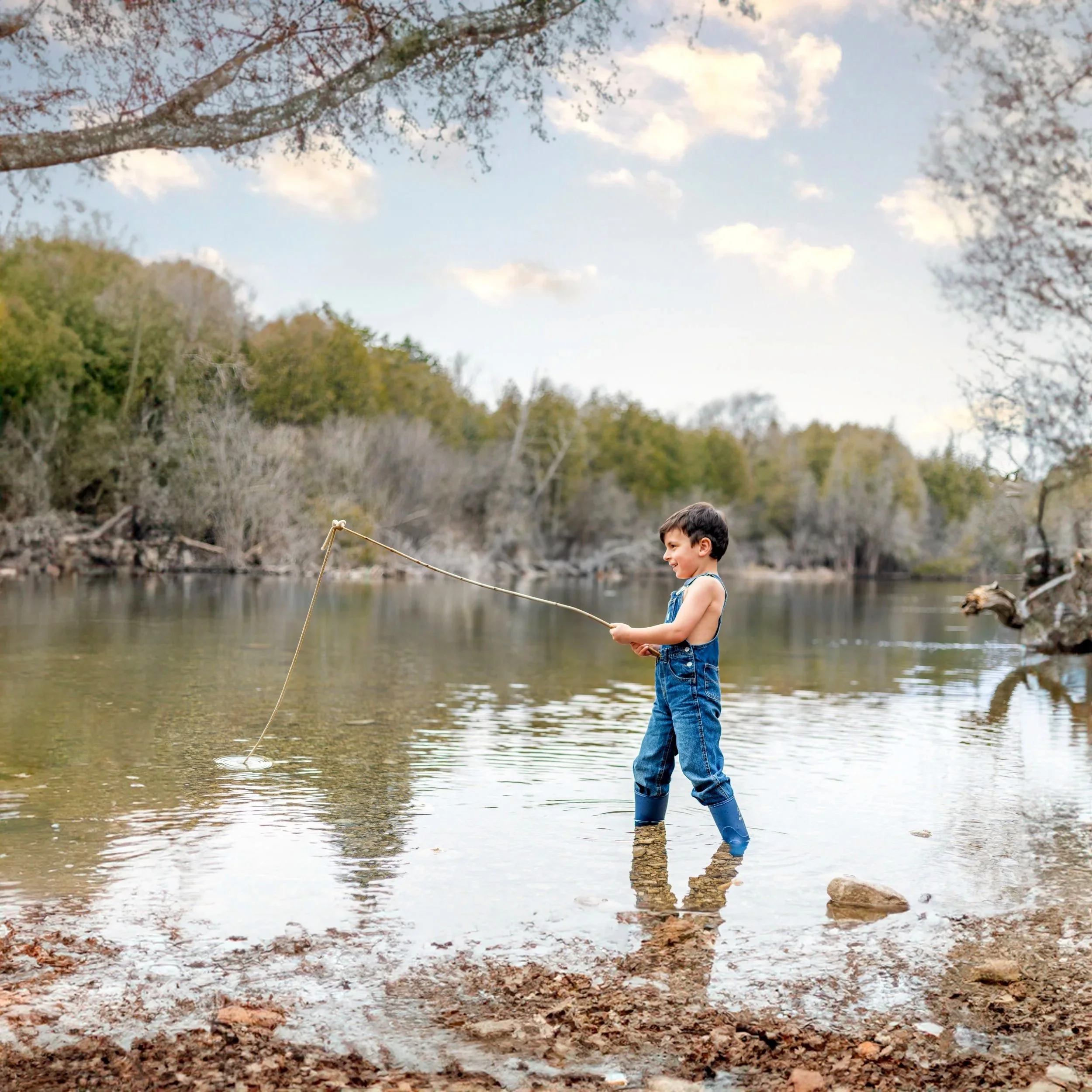 A young boy in overalls and rain boots fishing in a river surrounded by trees and a partly cloudy sky.