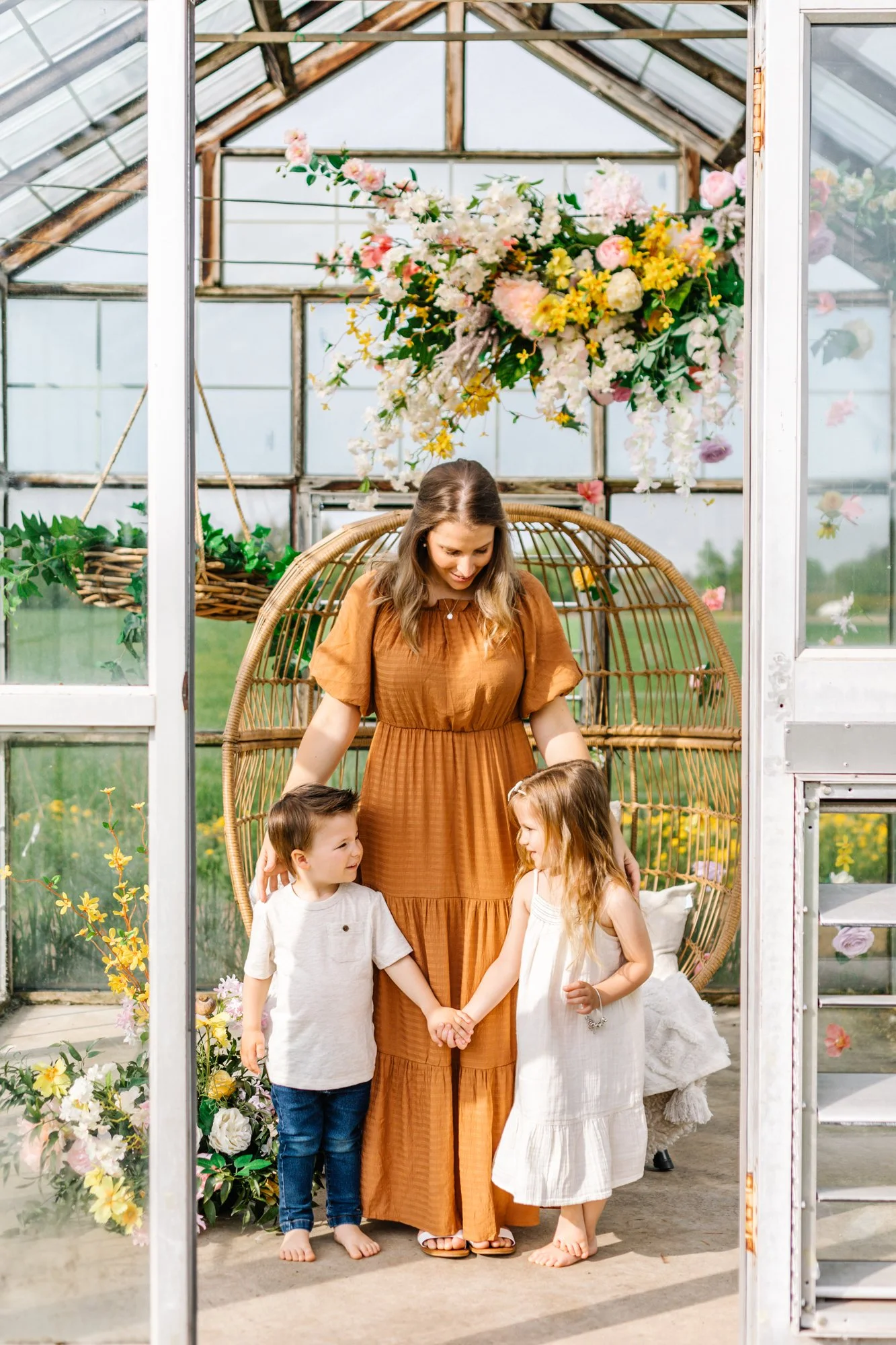 A woman in a brown dress stands with two young children in a greenhouse. The children hold hands, and there are colorful flowers and hanging plants in the background.