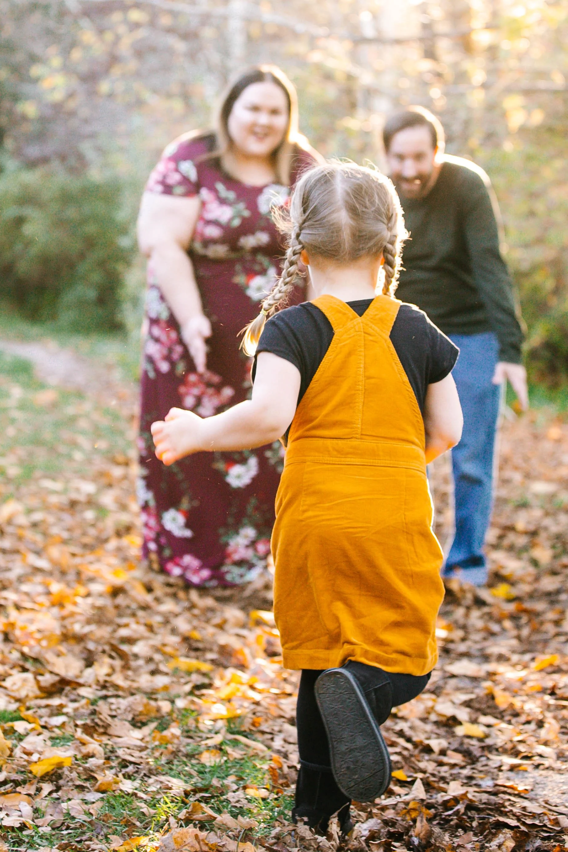 A child in a yellow dress running toward two adults in an autumn setting with fallen leaves and trees.