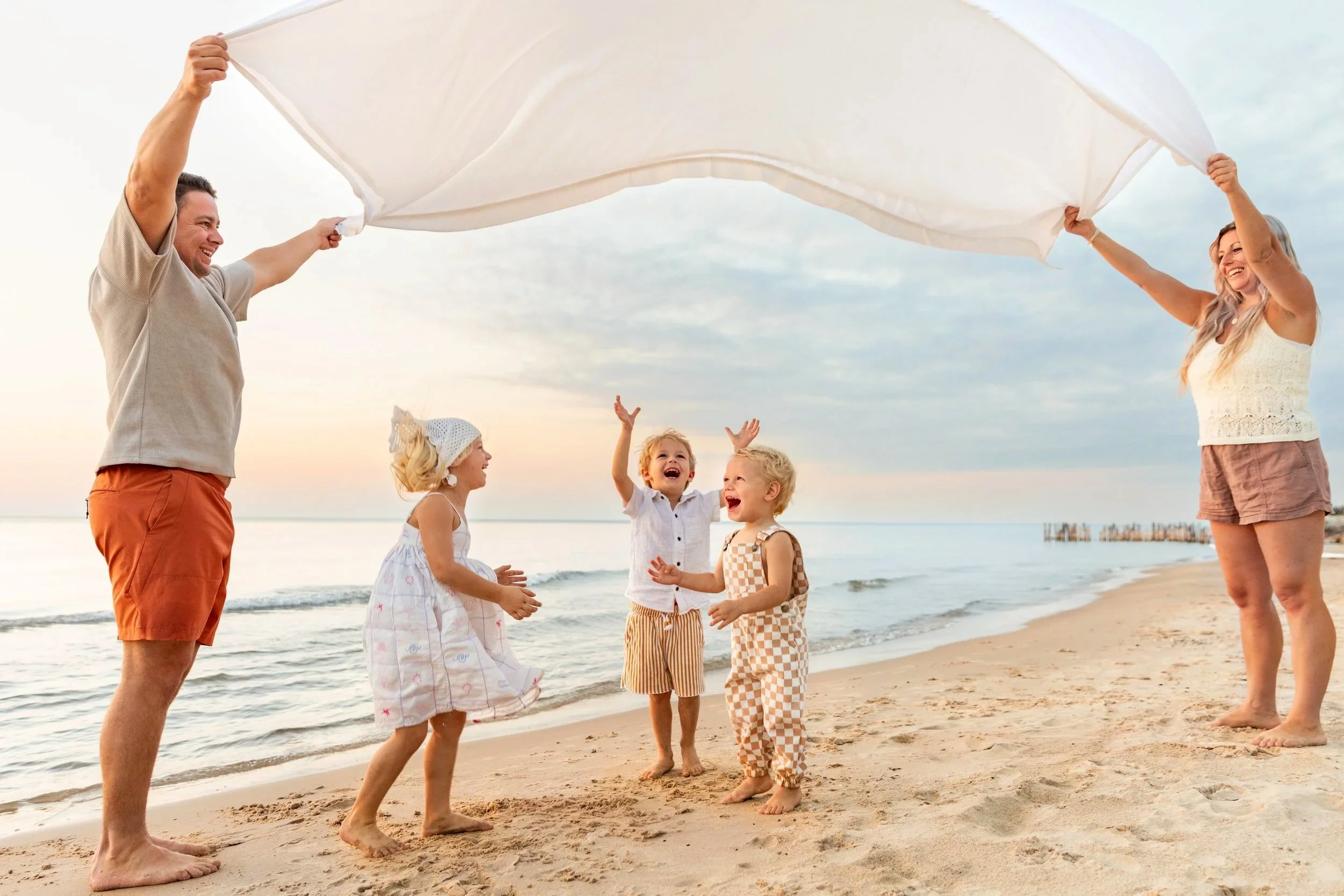 Family playing on the beach with a large white sheet, children and adults smiling and laughing at sunset.