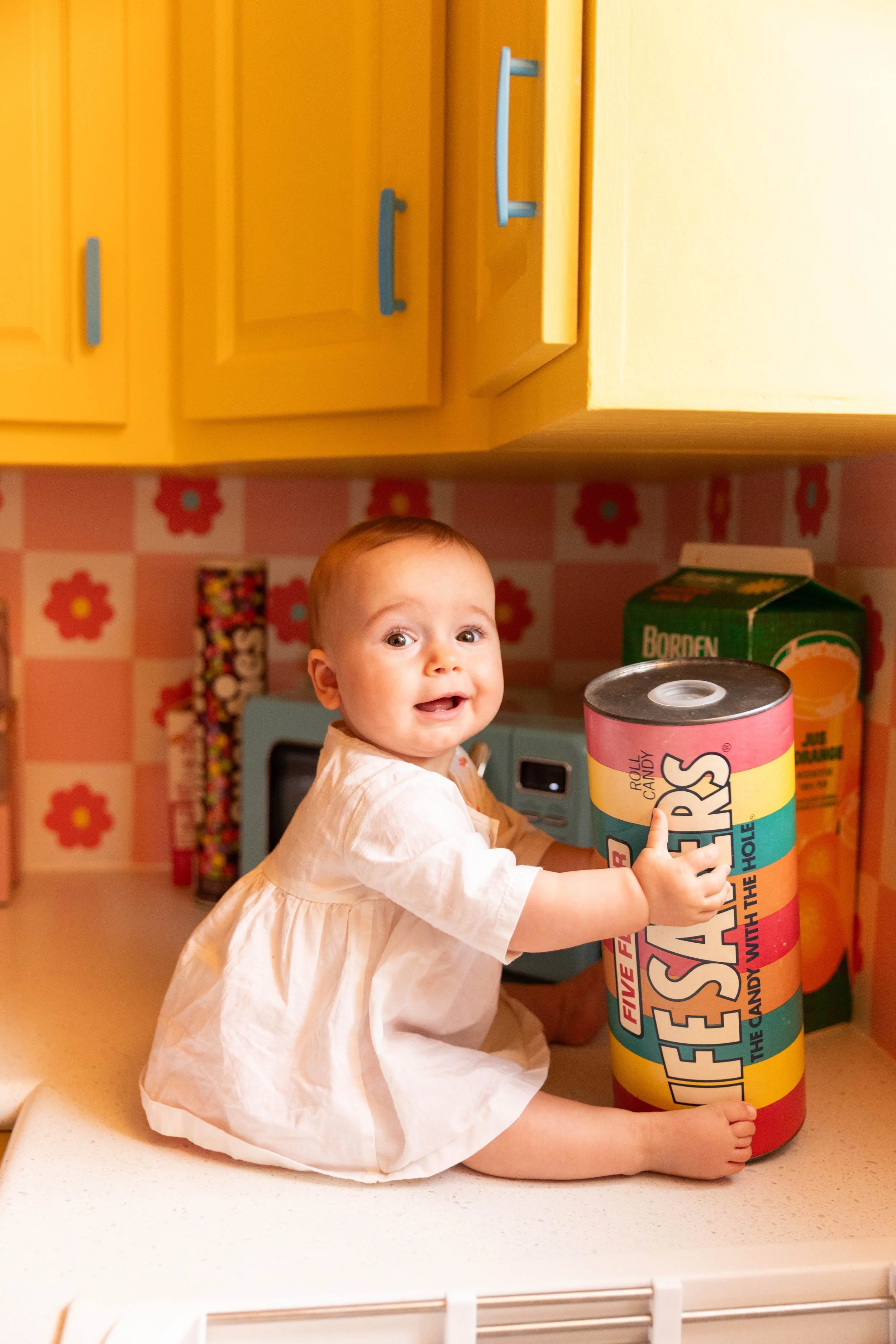 A toddler girl sitting on a kitchen counter, holding a large cylindrical container of colorful fireworks, with a smile, surrounded by kitchen appliances and yellow cabinets.