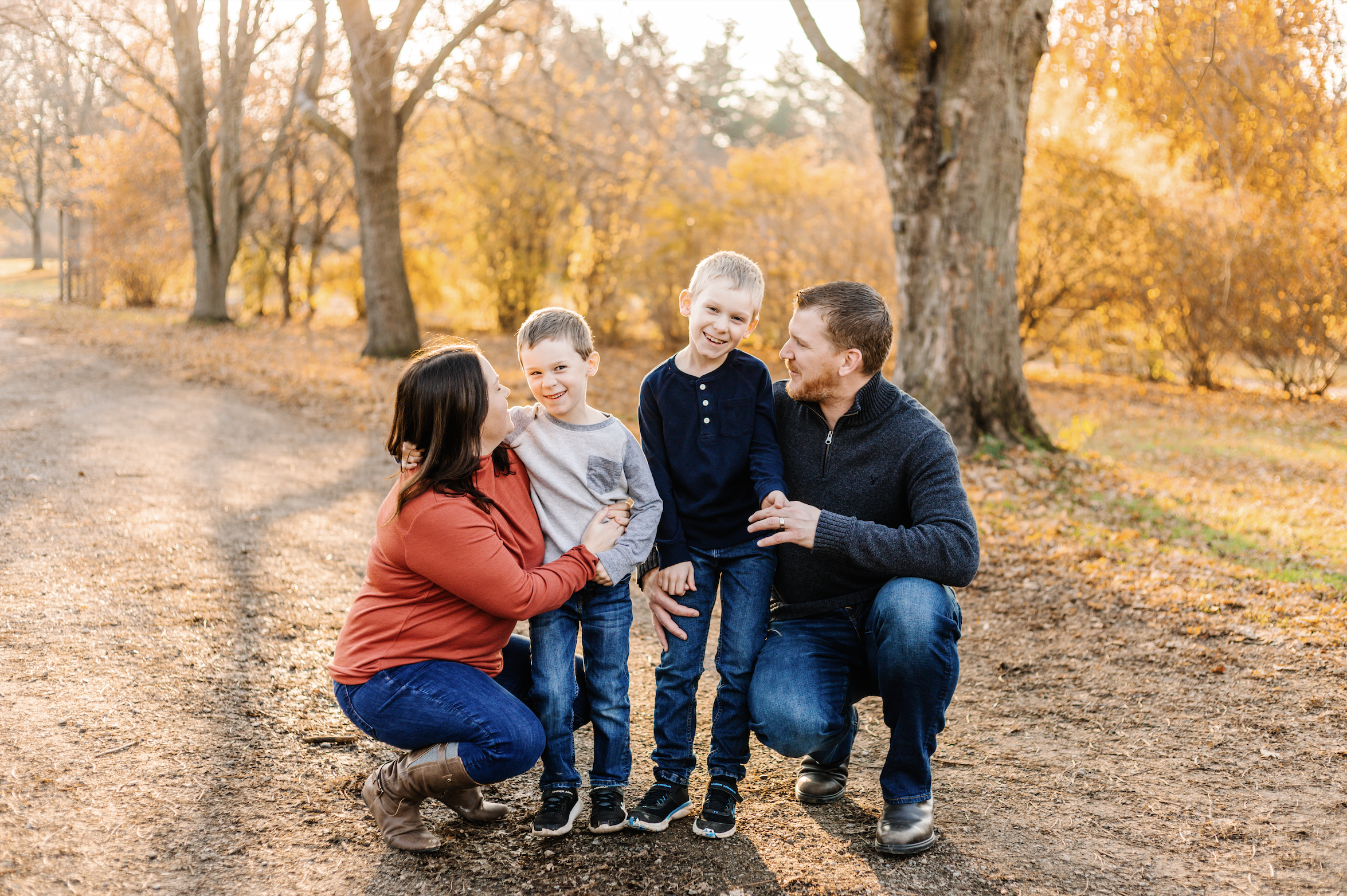 Family of four kneeling and smiling in a park during autumn, with trees and fallen leaves in the background.