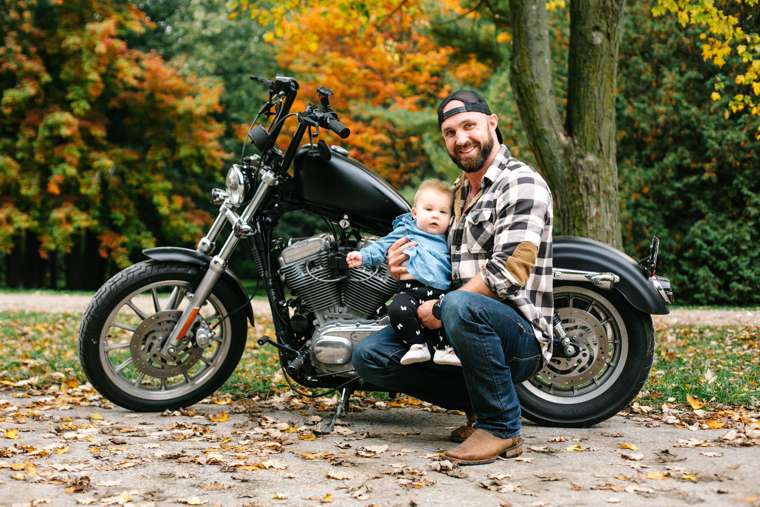 Father holding baby next to a black motorcycle in a park with autumn foliage.