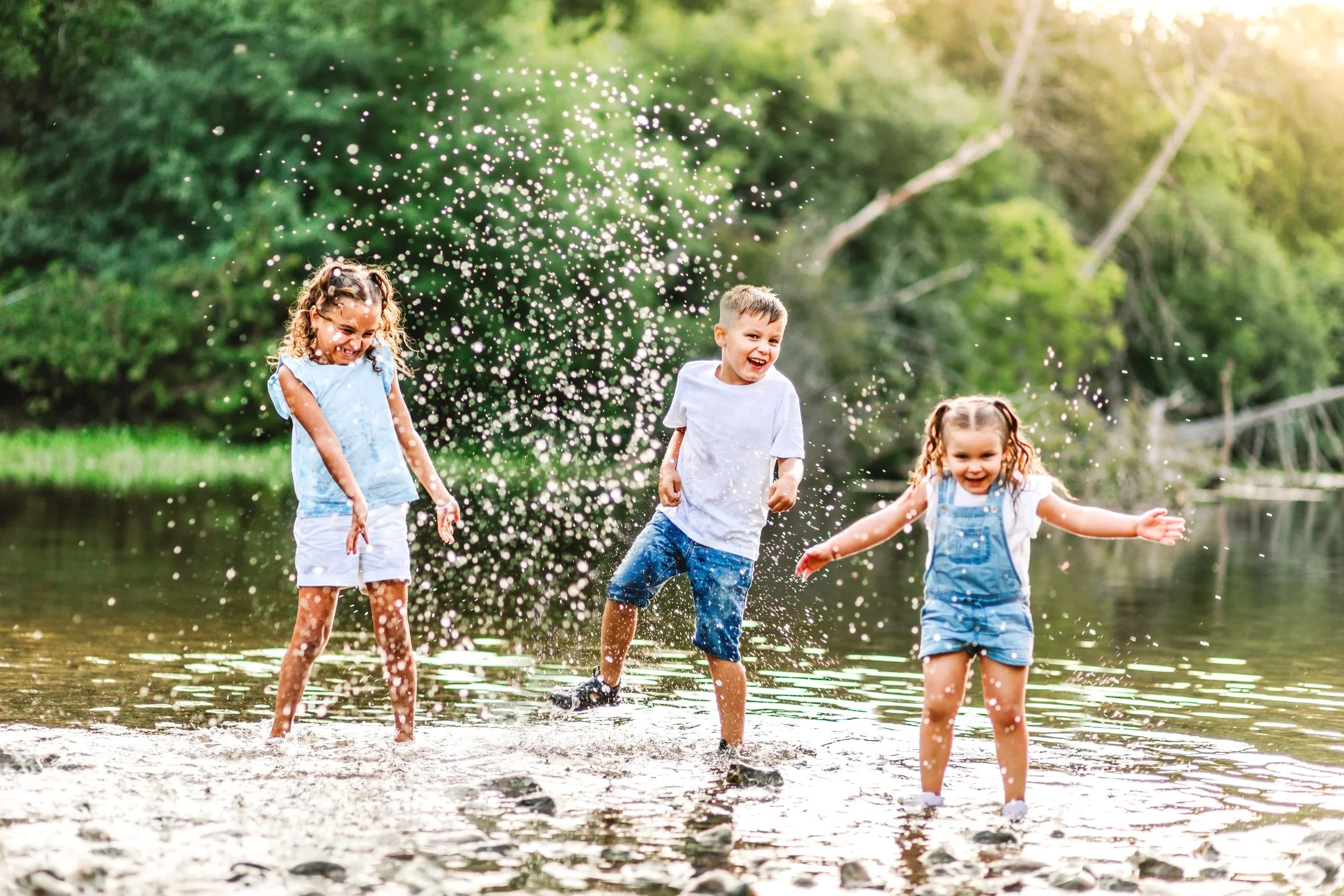 Three children playing and splashing water in a shallow river with greenery in the background.