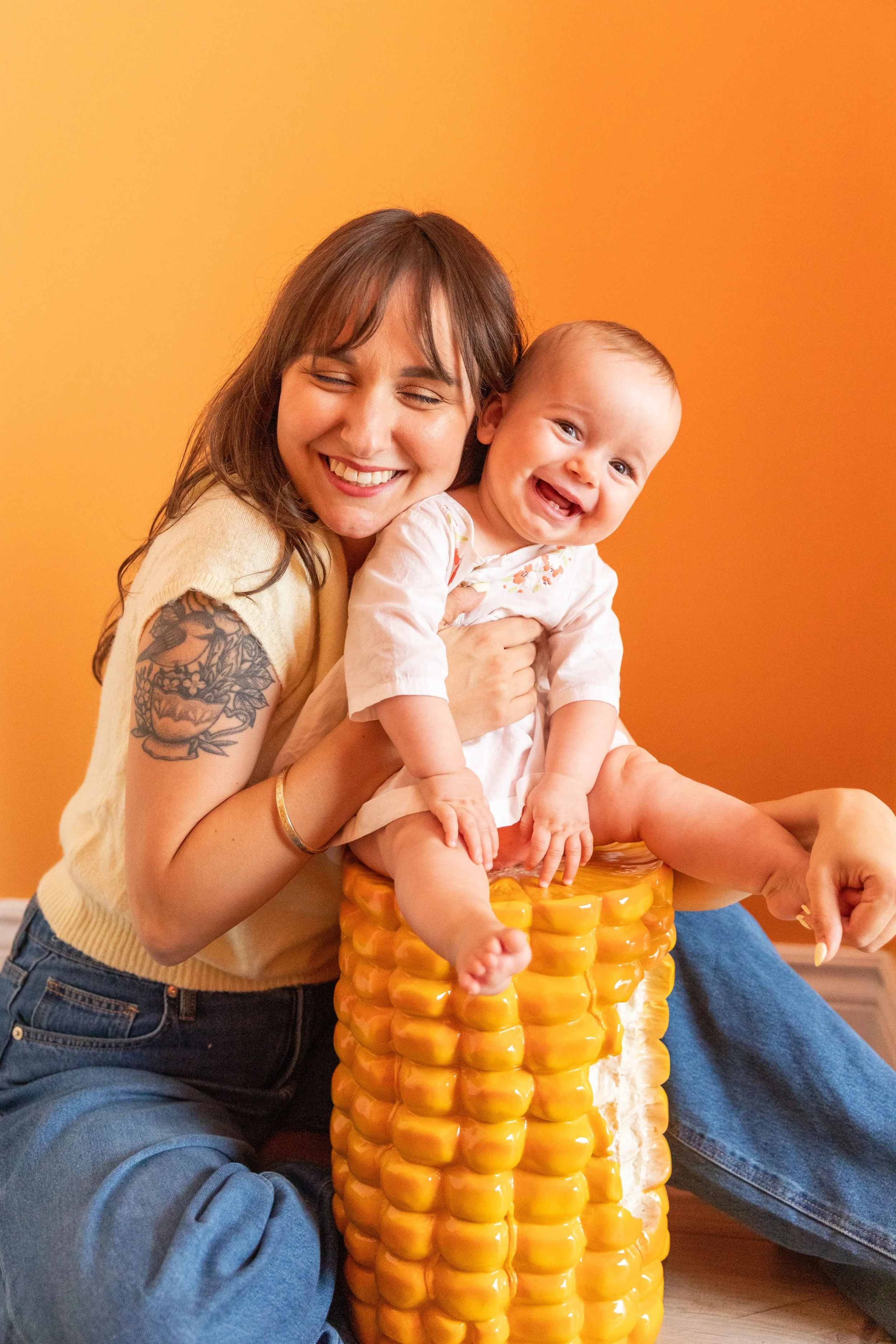 A woman with shoulder-length brown hair, wearing a cream sleeveless top, holding a smiling baby on a yellow plastic corn cob-shaped chair against an orange background.