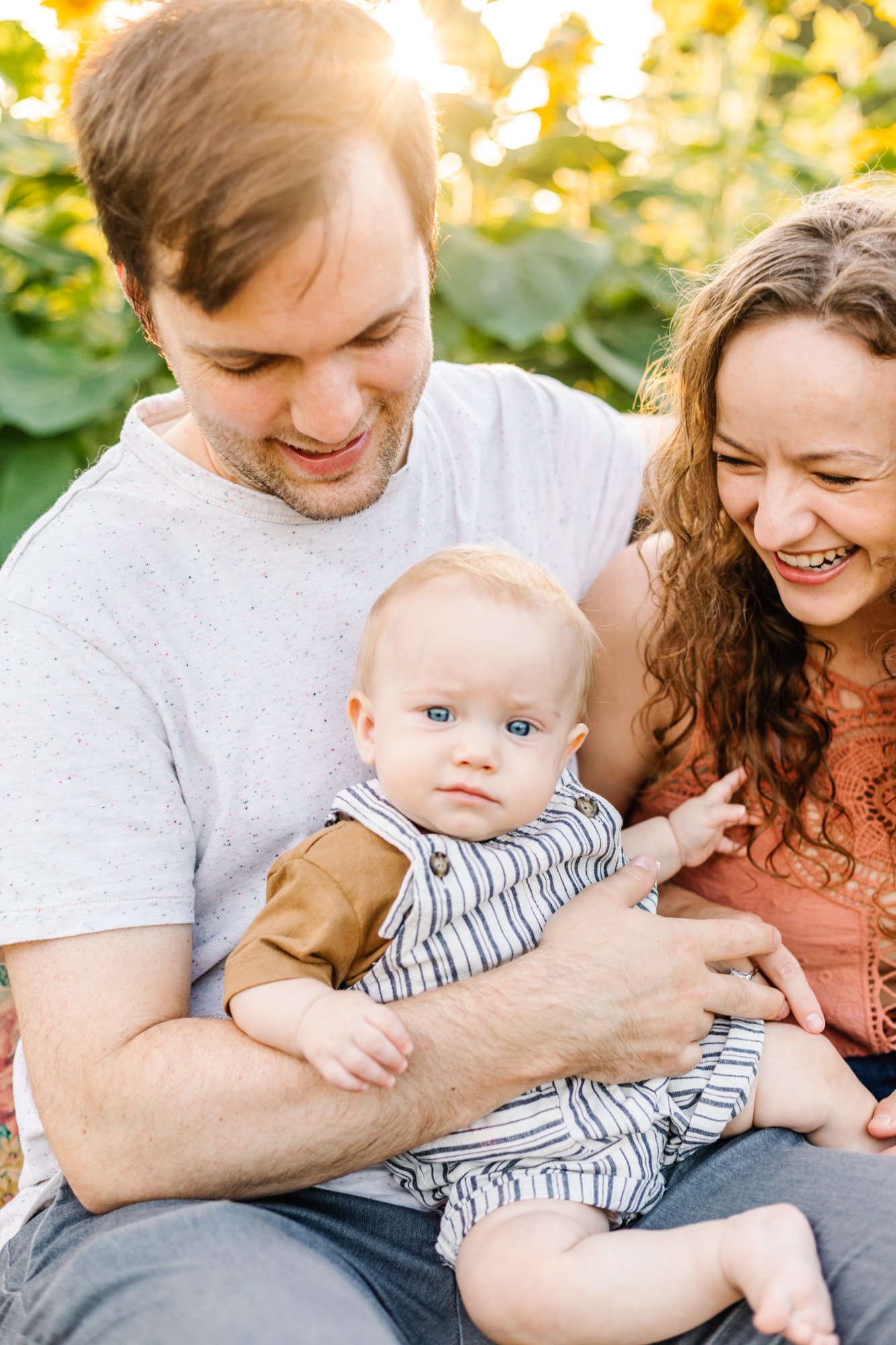 A family with a baby outdoors, surrounded by sunflowers, during a sunny day.