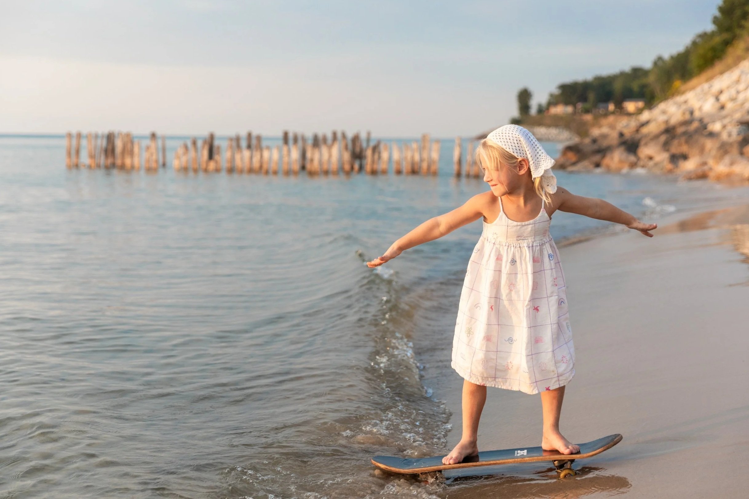A young girl wearing a white dress and a white sun hat balancing on a skateboard at the shoreline, with her arms outstretched and water gently lapping at her feet, in a beach setting with a rocky coast and wooden breakwater in the background.