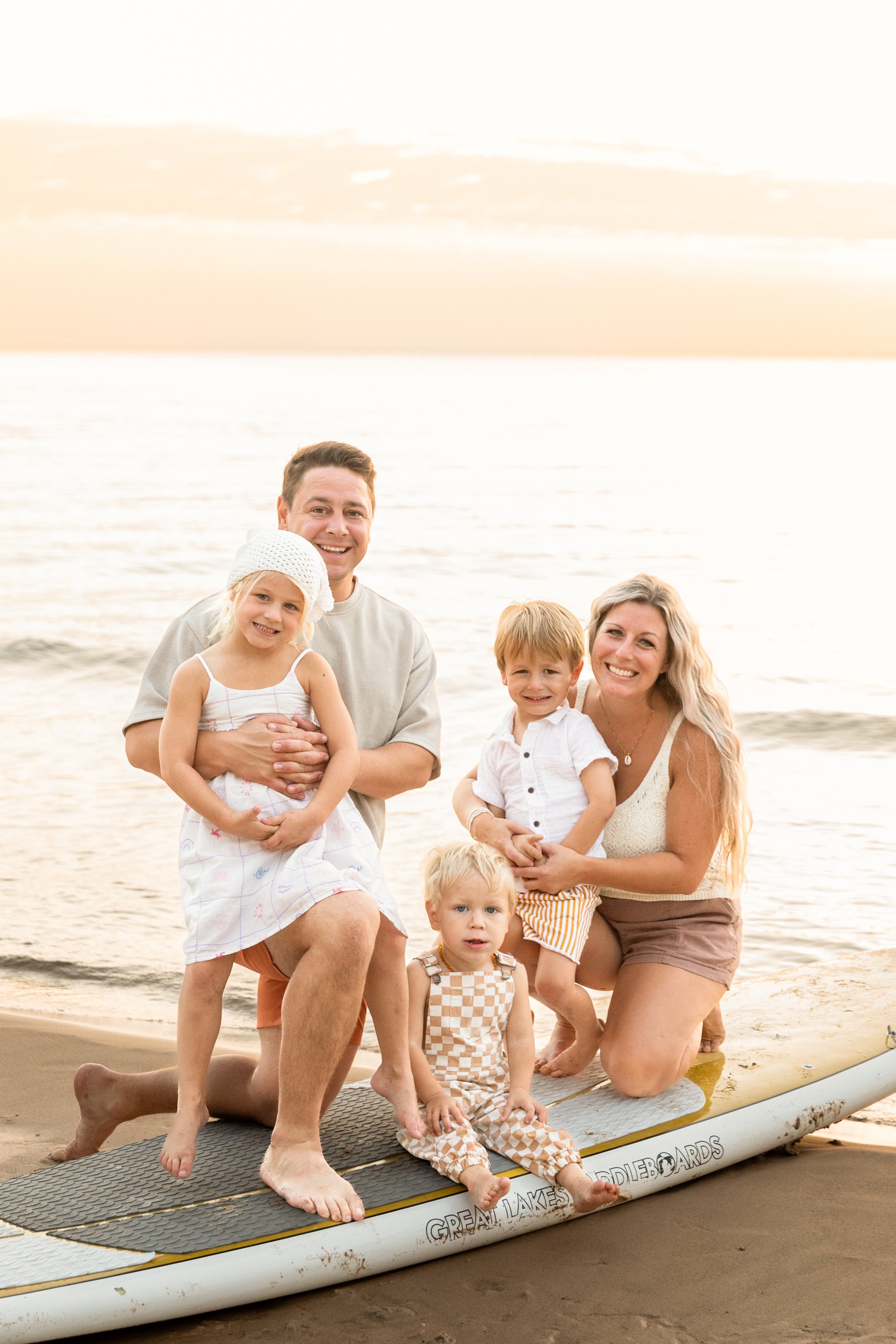 A family of five posing on a surfboard on the beach during sunset.