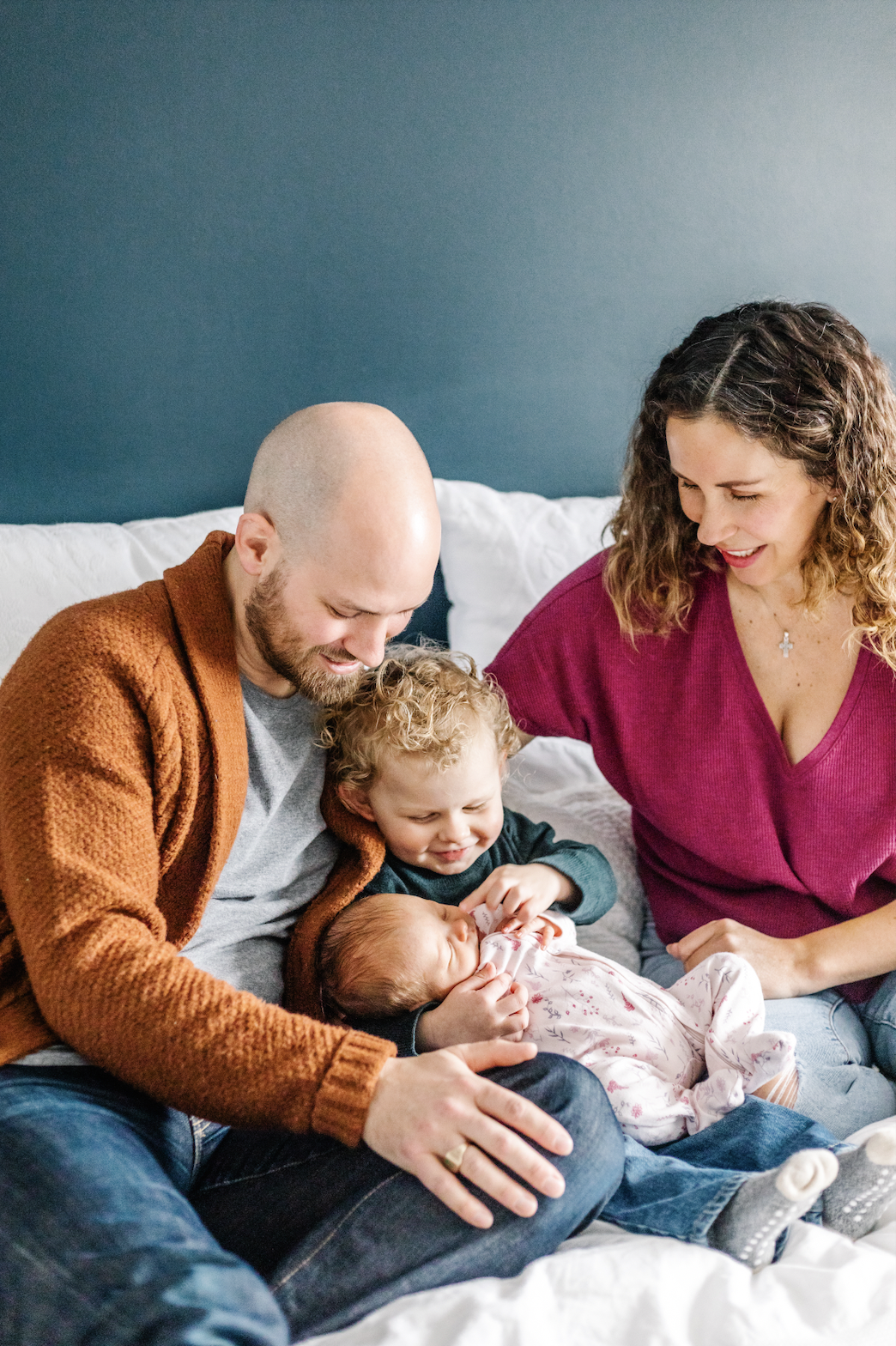 A family of four sitting on a bed, smiling at a newborn baby. A child reaches to touch the baby, with a man and woman sitting on either side.