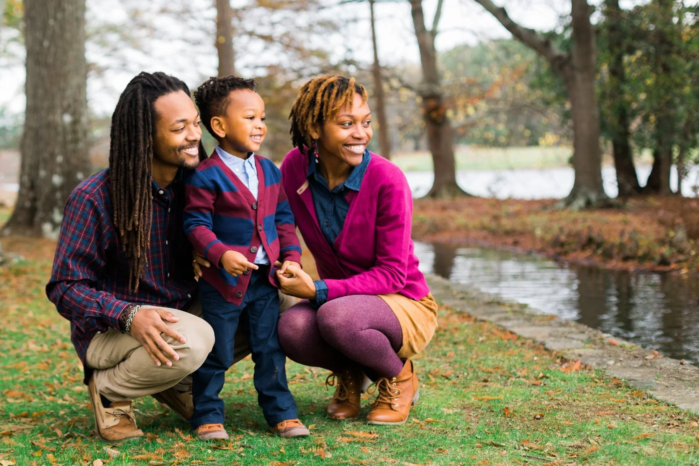 A family of three, including a man, woman, and young boy, crouching and smiling by a pond in a park during the fall season. The boy wears a colorful cardigan, and the adults are dressed in casual fall attire with jackets and boots.