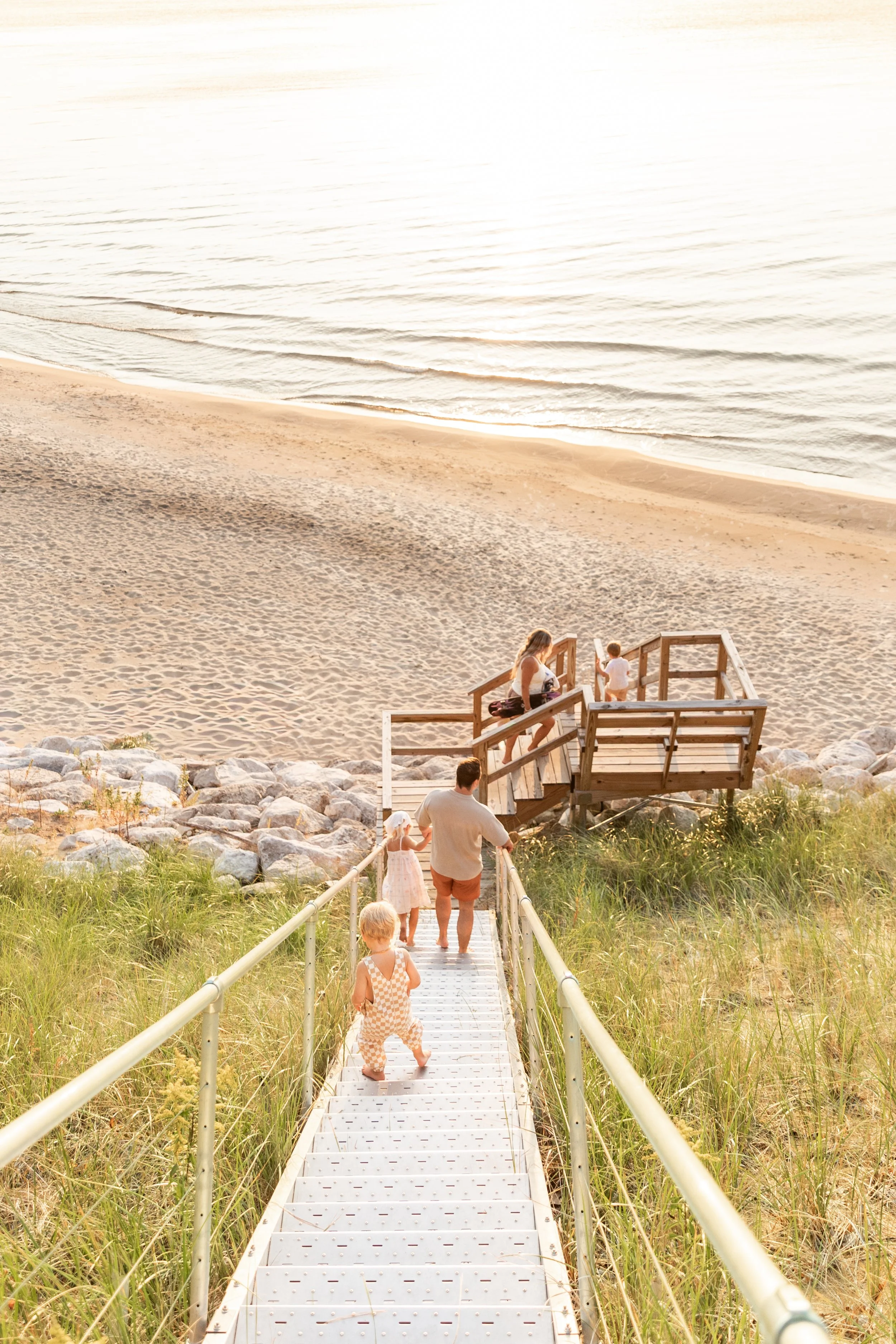 Family walking down a metal staircase towards the beach with sand and water beyond.