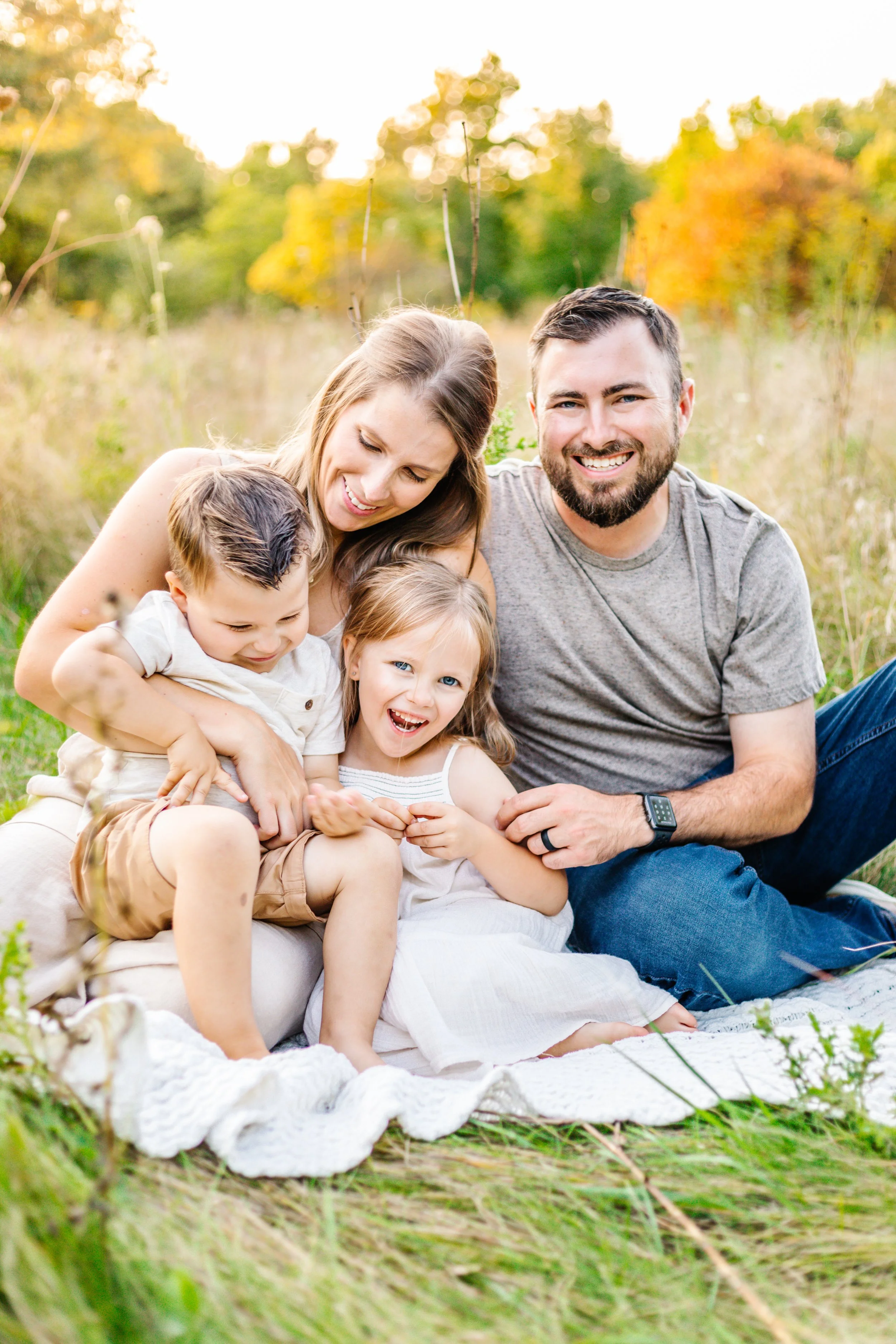 A family of four sitting on a blanket in a grassy field, with a man, woman, and two children smiling and interacting playfully in a sunny outdoor setting.