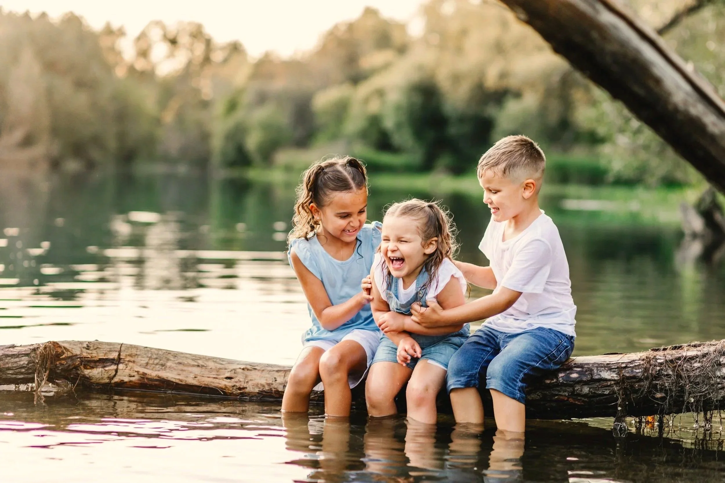 Three children sitting on a log in a lake, laughing and playing. The background features trees and water.