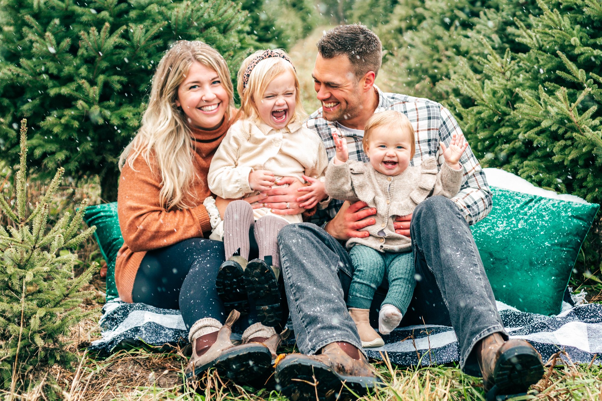 A family of four sitting on a blanket among evergreen trees, smiling and laughing. Two adults and two children, wearing casual winter clothing.