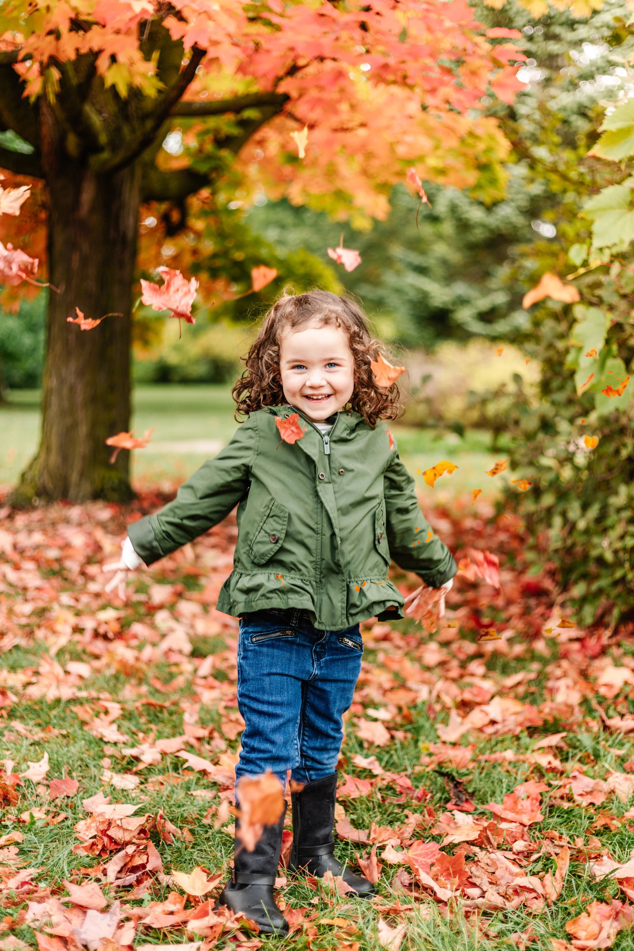 Smiling child in green jacket playing in autumn leaves with colorful trees in the background.