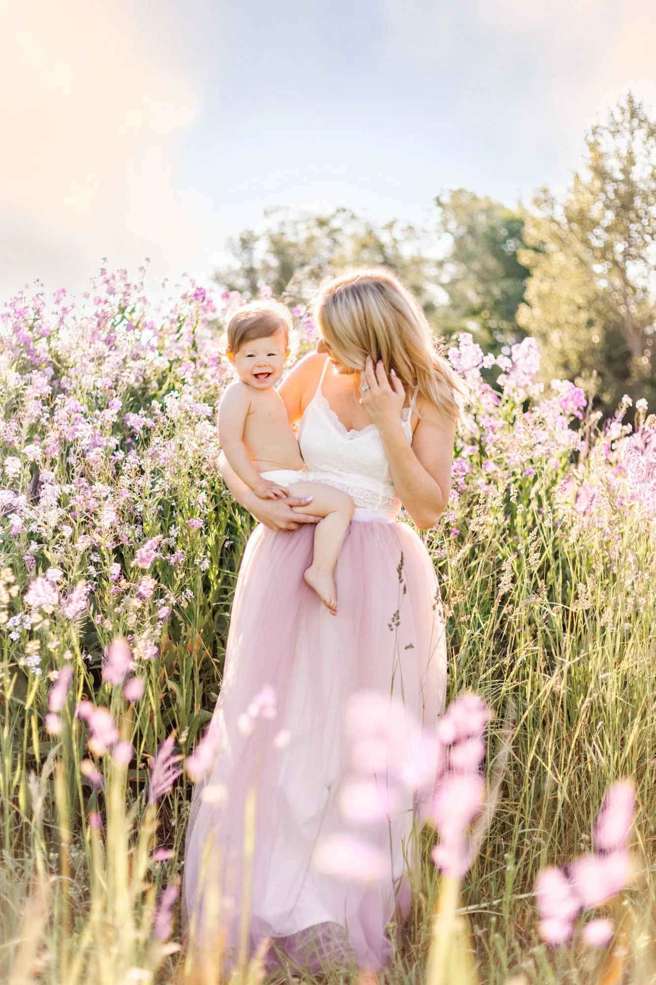 A woman holding a happy baby in a field of purple flowers, wearing a white top and a pink tulle skirt.