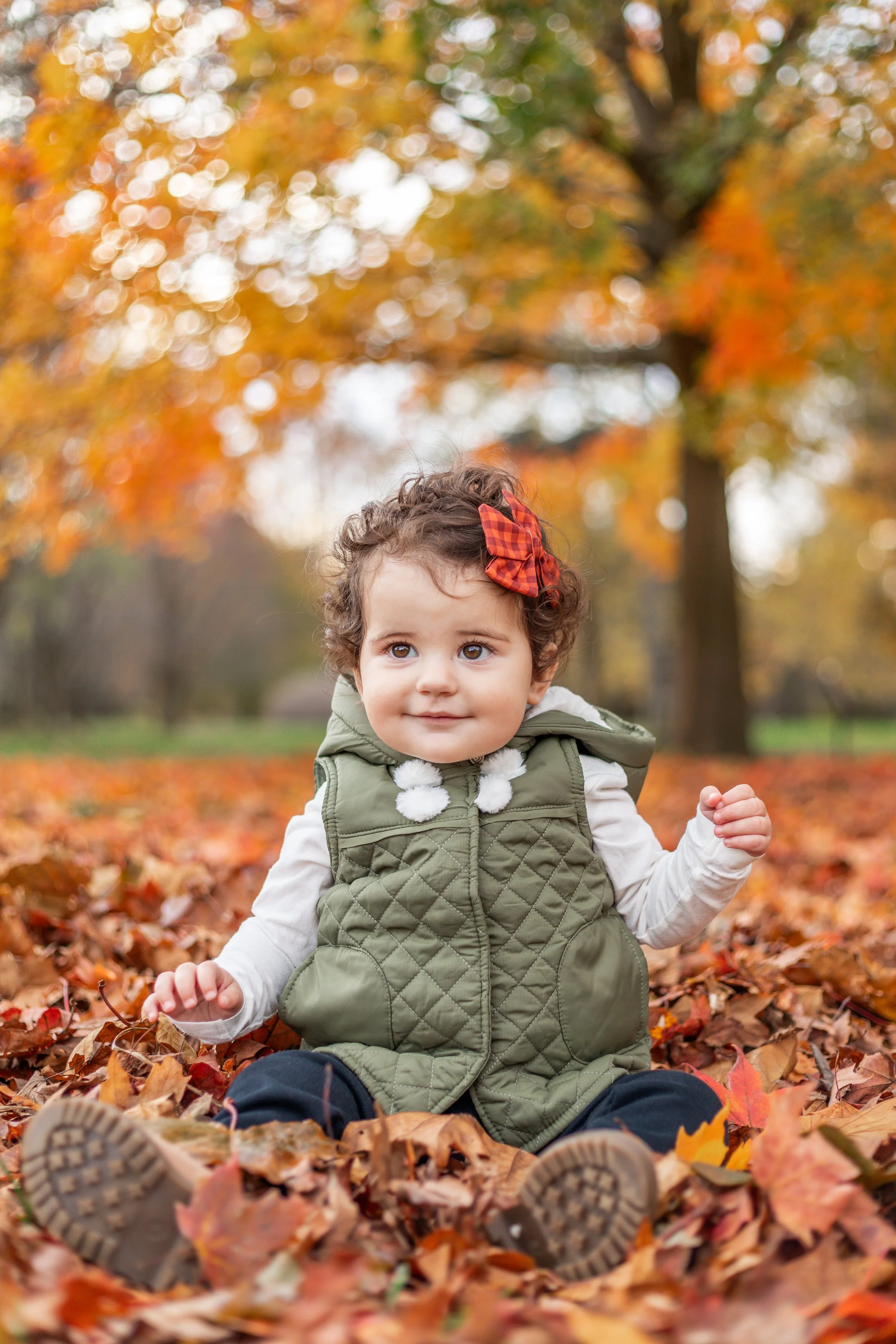 A young child with curly hair and a red plaid bow sits on autumn leaves, wearing a green quilted vest. Background has colorful fall foliage.