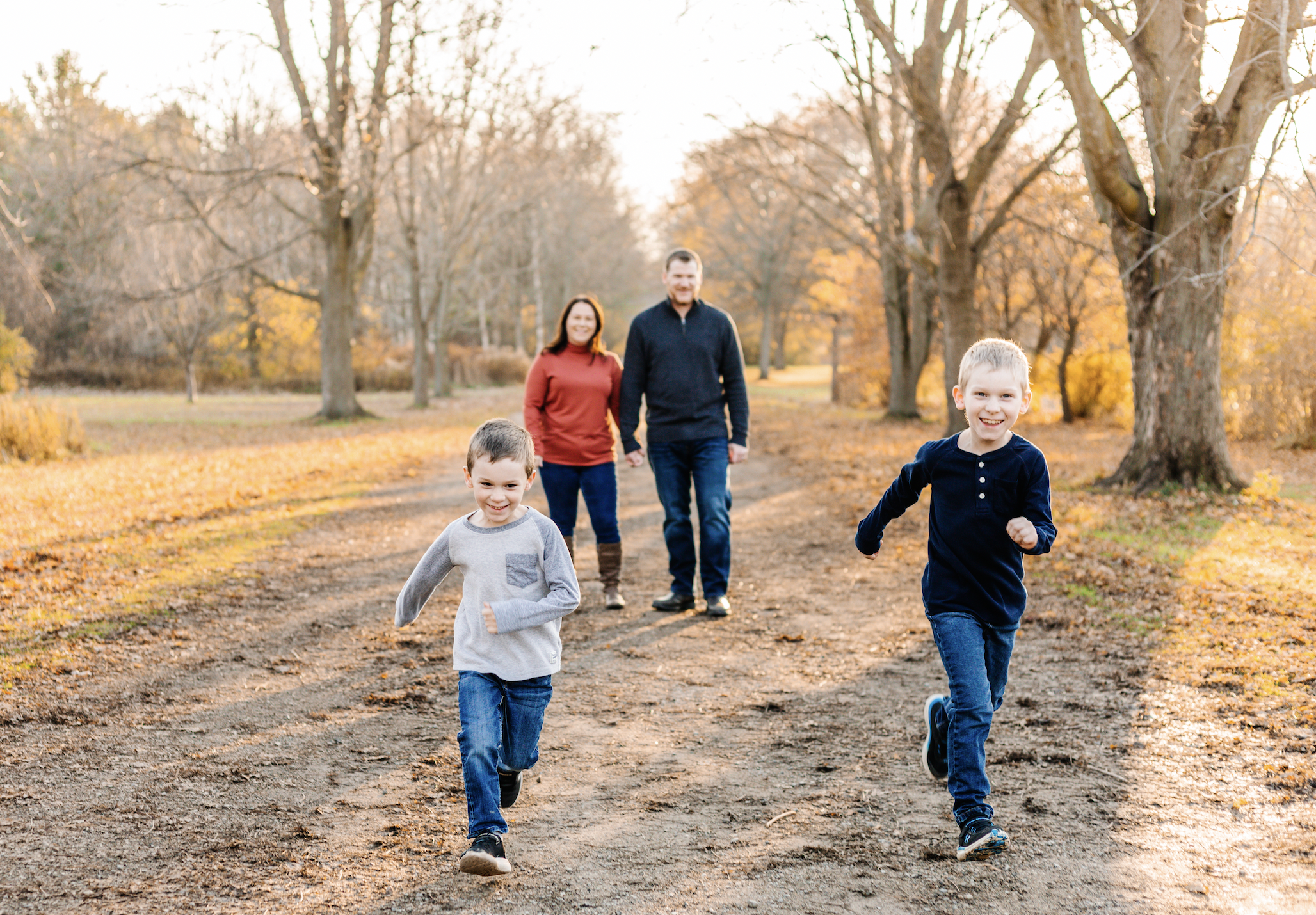 Two children running on a dirt path with two adults walking behind them in a park with autumn foliage.