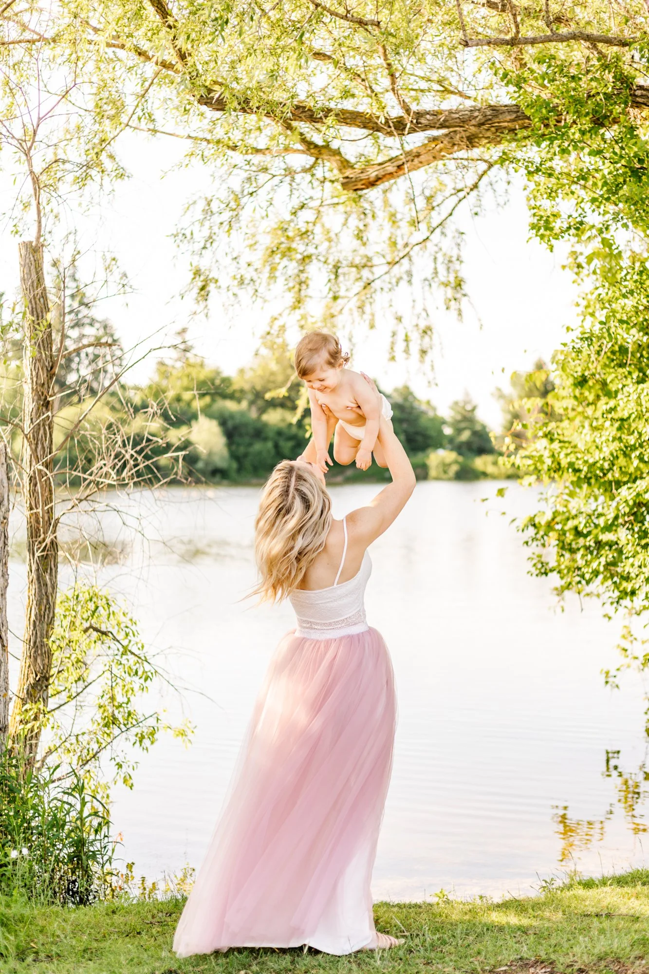 A woman in a white top and pink skirt holds a baby above her head near a lake, surrounded by greenery.
