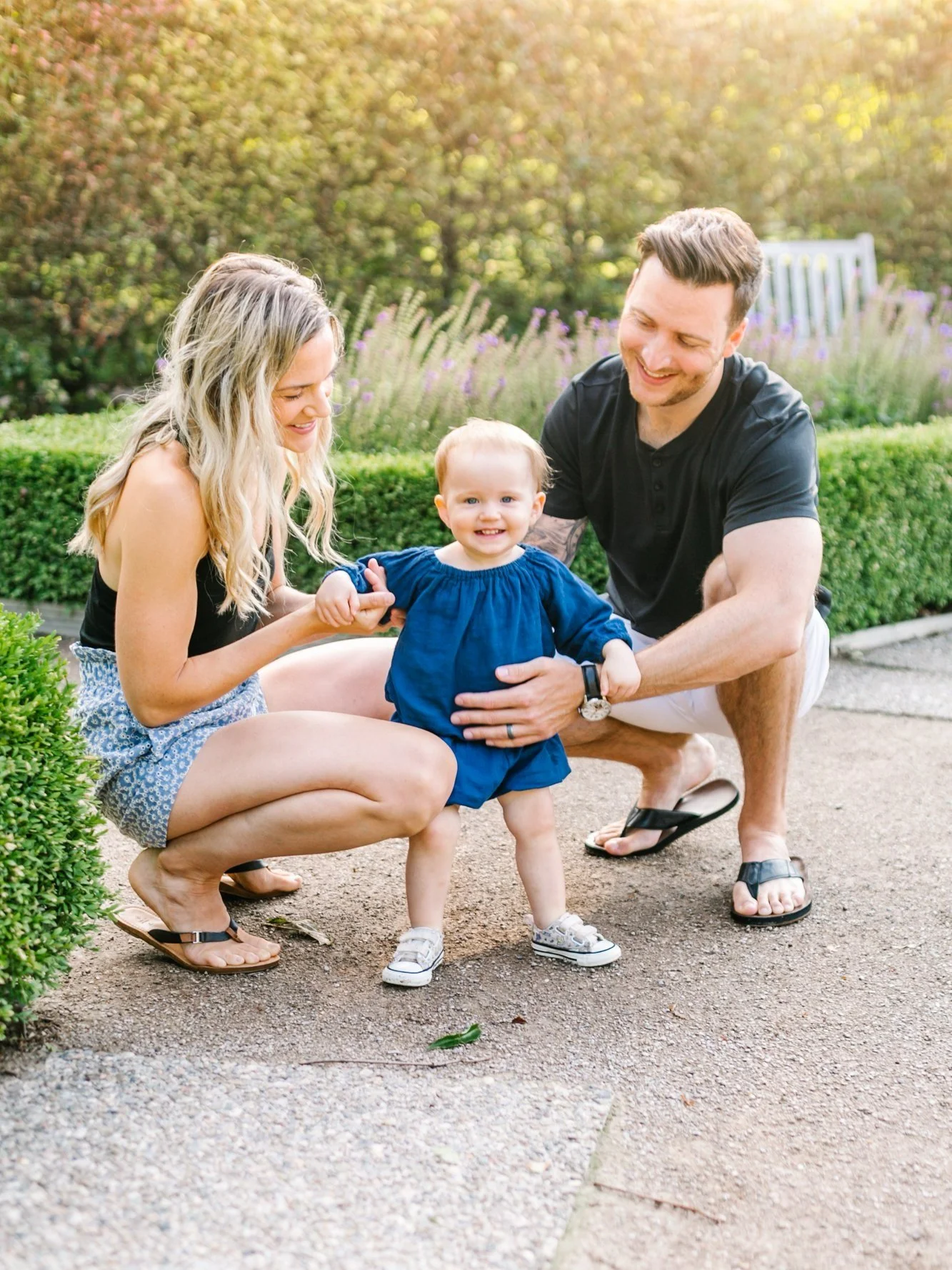 A woman and man crouching, smiling at a toddler in a blue dress standing between them in a sunny garden setting.