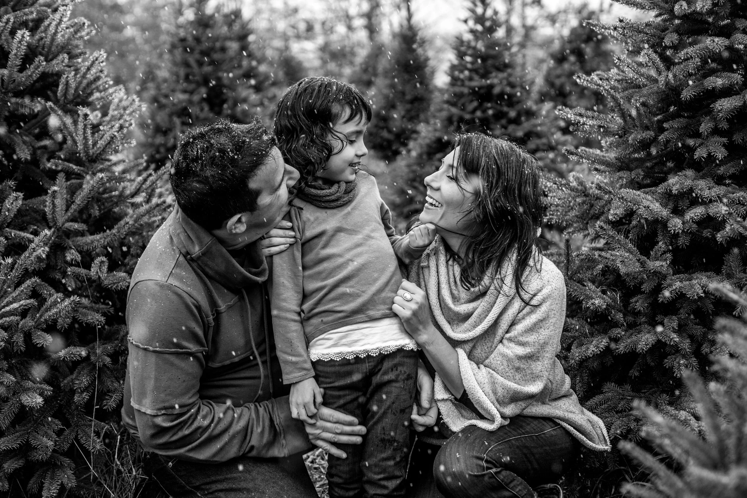 Black and white photo of a joyful family in winter clothing, surrounded by evergreen trees, smiling and embracing each other, with light snowfall around them.
