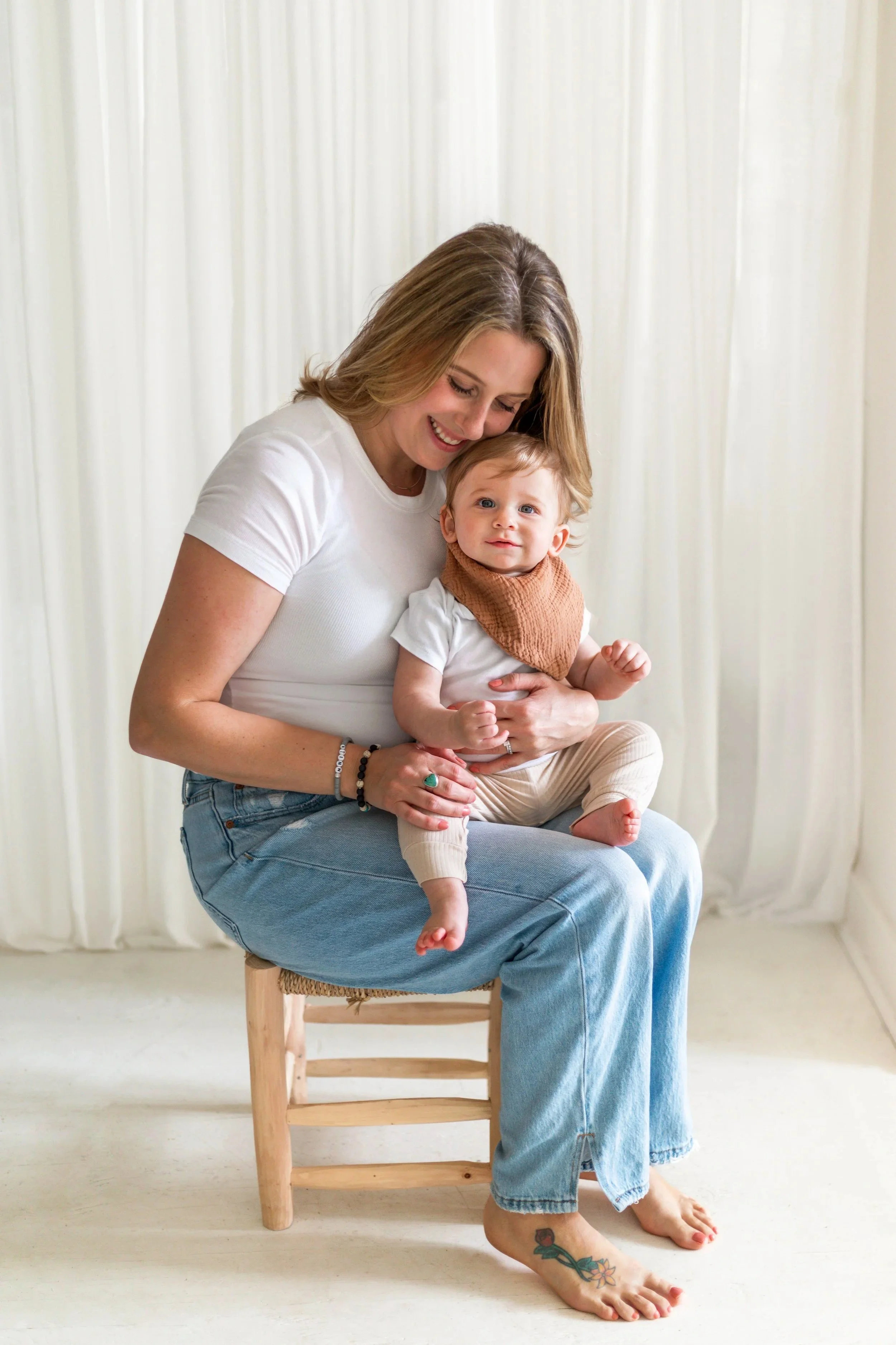 A smiling woman sitting on a wooden chair holding a baby on her lap. The woman has blonde hair, is wearing a white t-shirt and blue jeans, and has a tattoo on her left foot. The baby has light brown hair, is wearing a white shirt, beige pants, and a 