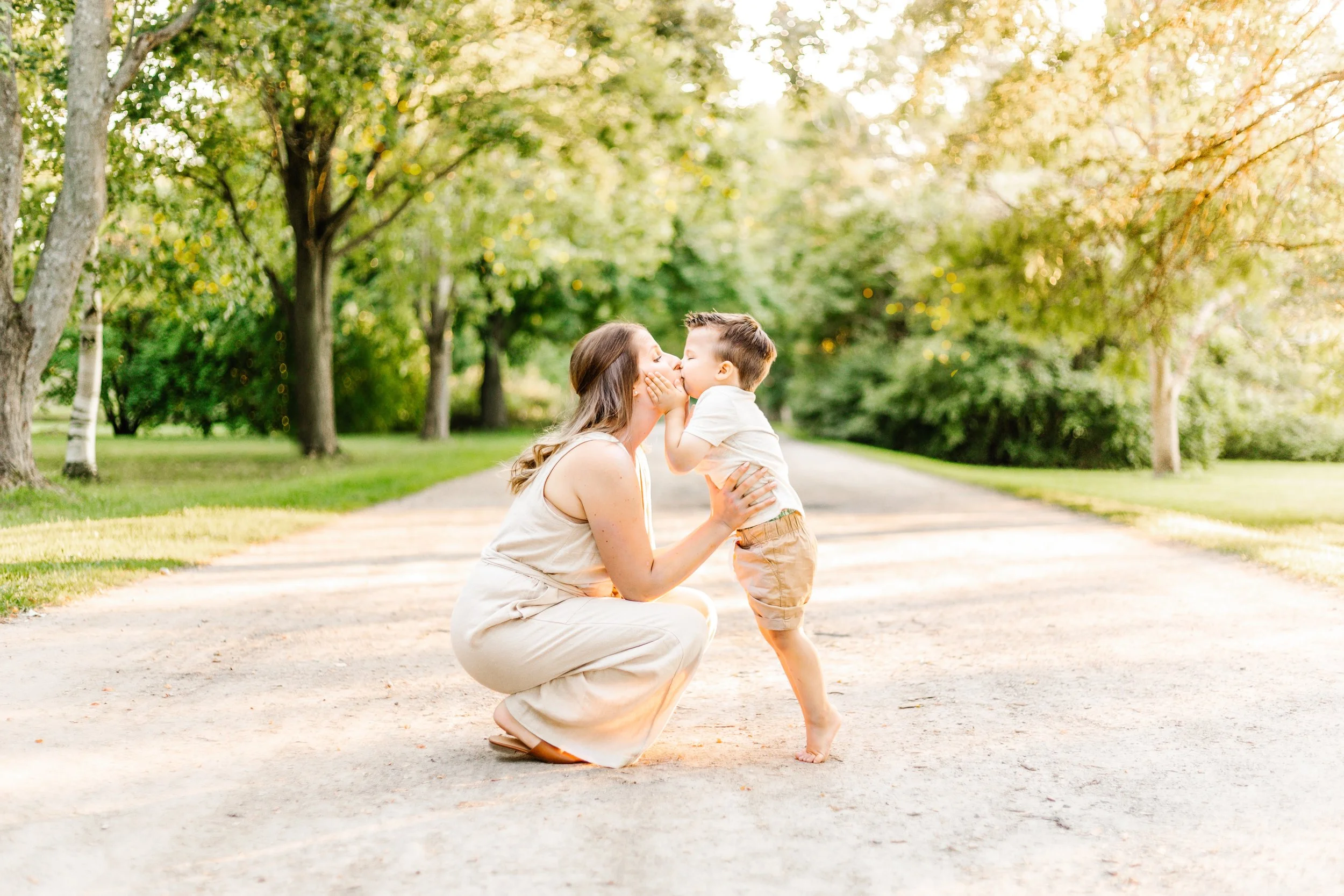 A woman kneeling on a dirt path kissing a young boy standing in front of her, surrounded by trees in a park.
