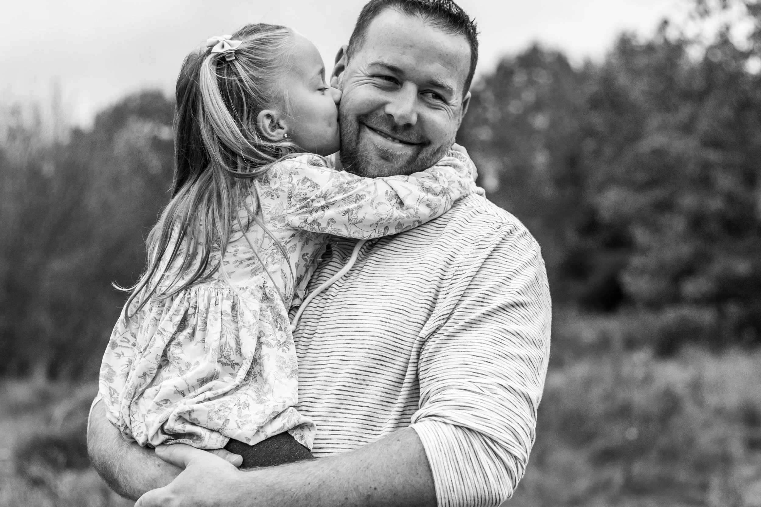 Black and white photo of a little girl kissing a smiling man on the cheek, outdoors.