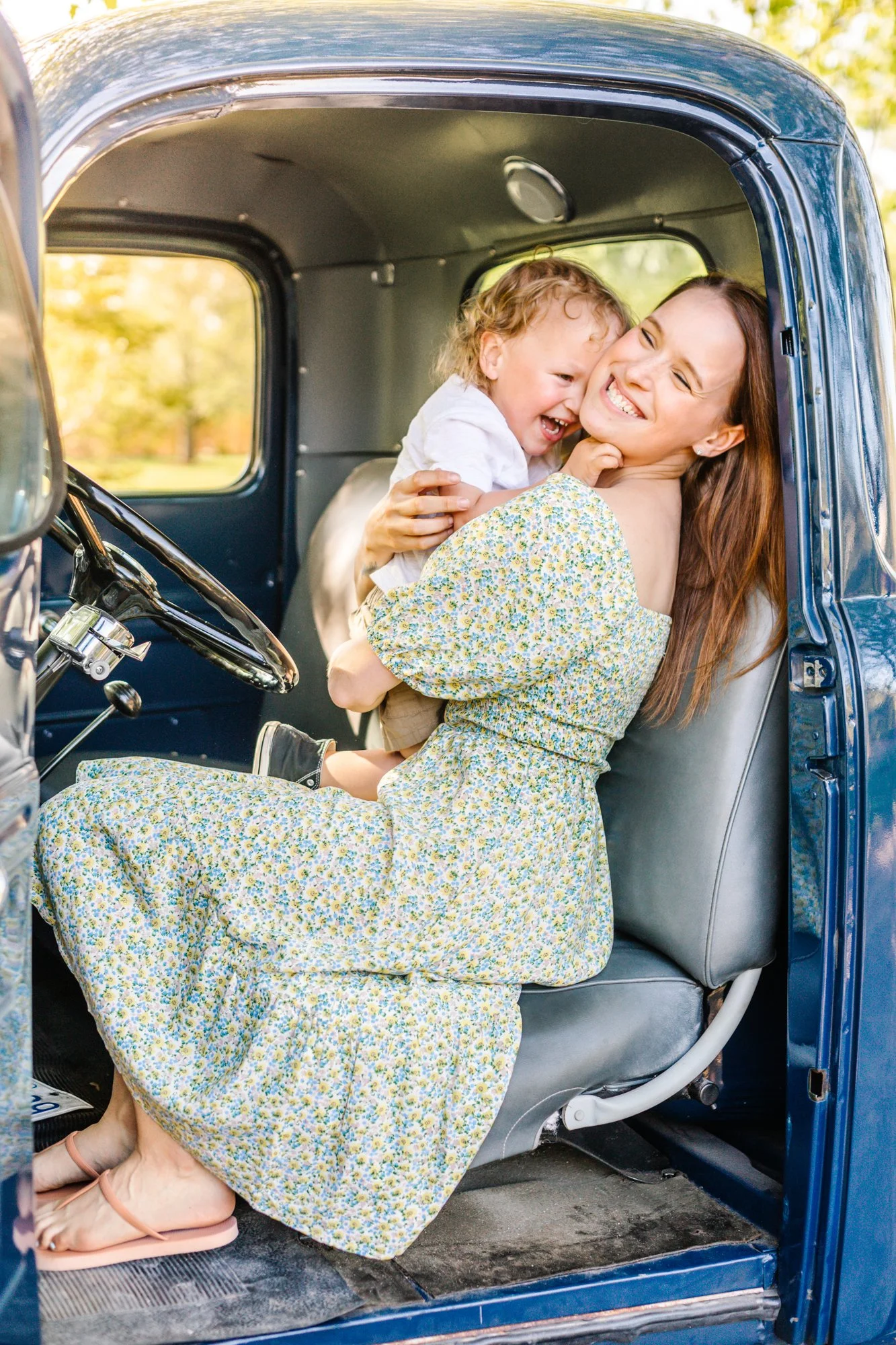 A woman and a young child sitting inside a vintage blue truck, smiling and laughing together during daytime.